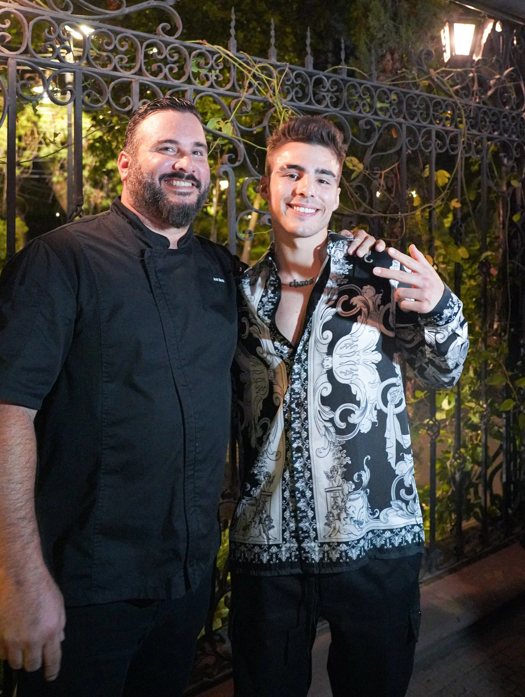 Two men are standing outdoors at night, smiling for a photo. The man on the left has a beard and short hair, wearing a black chef's coat. The man on the right has short dark hair, wearing a patterned black and white shirt, and making a peace sign wit