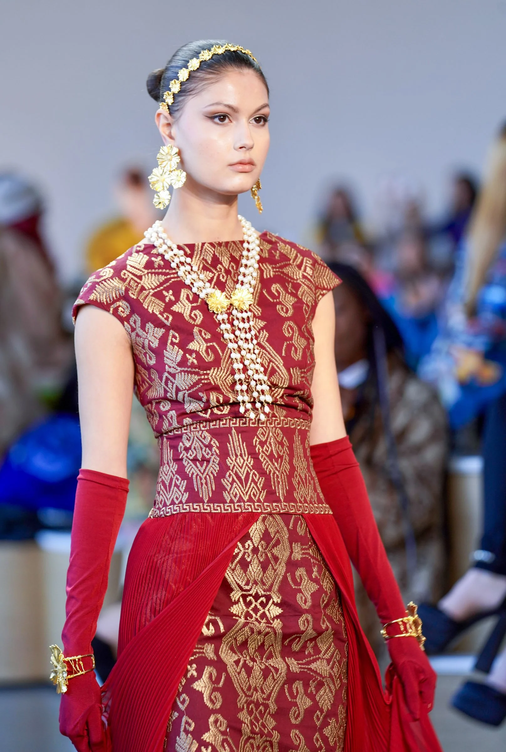A woman with dark hair styled in an elegant bun wearing a traditional red and gold patterned dress, adorned with large floral earrings, multiple layered pearl necklaces, and gold accessories, walking in an indoor setting.