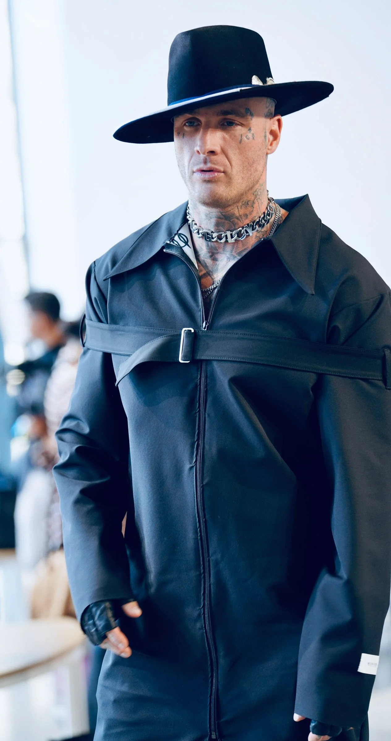 A man wearing a wide-brimmed black hat with a blue band, black leather jacket, multiple tattoos on his face and neck, and accessories including necklaces and rings, walking in an indoor setting.