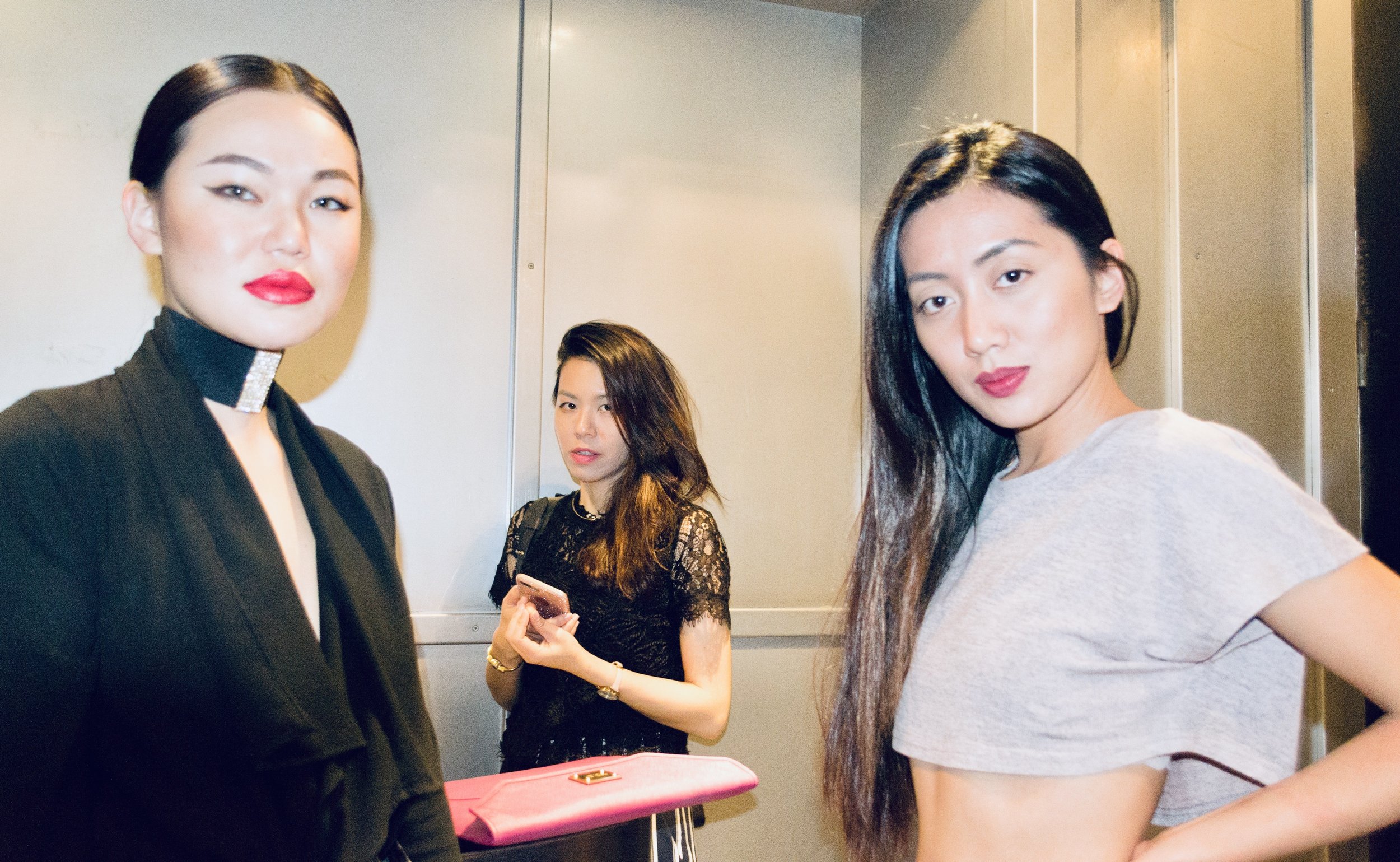 Four women in an elevator, two facing the camera and two looking away, one with bright red lipstick and a black outfit, another in a gray t-shirt, one with long dark hair holding a phone, and the third with a black lace dress.