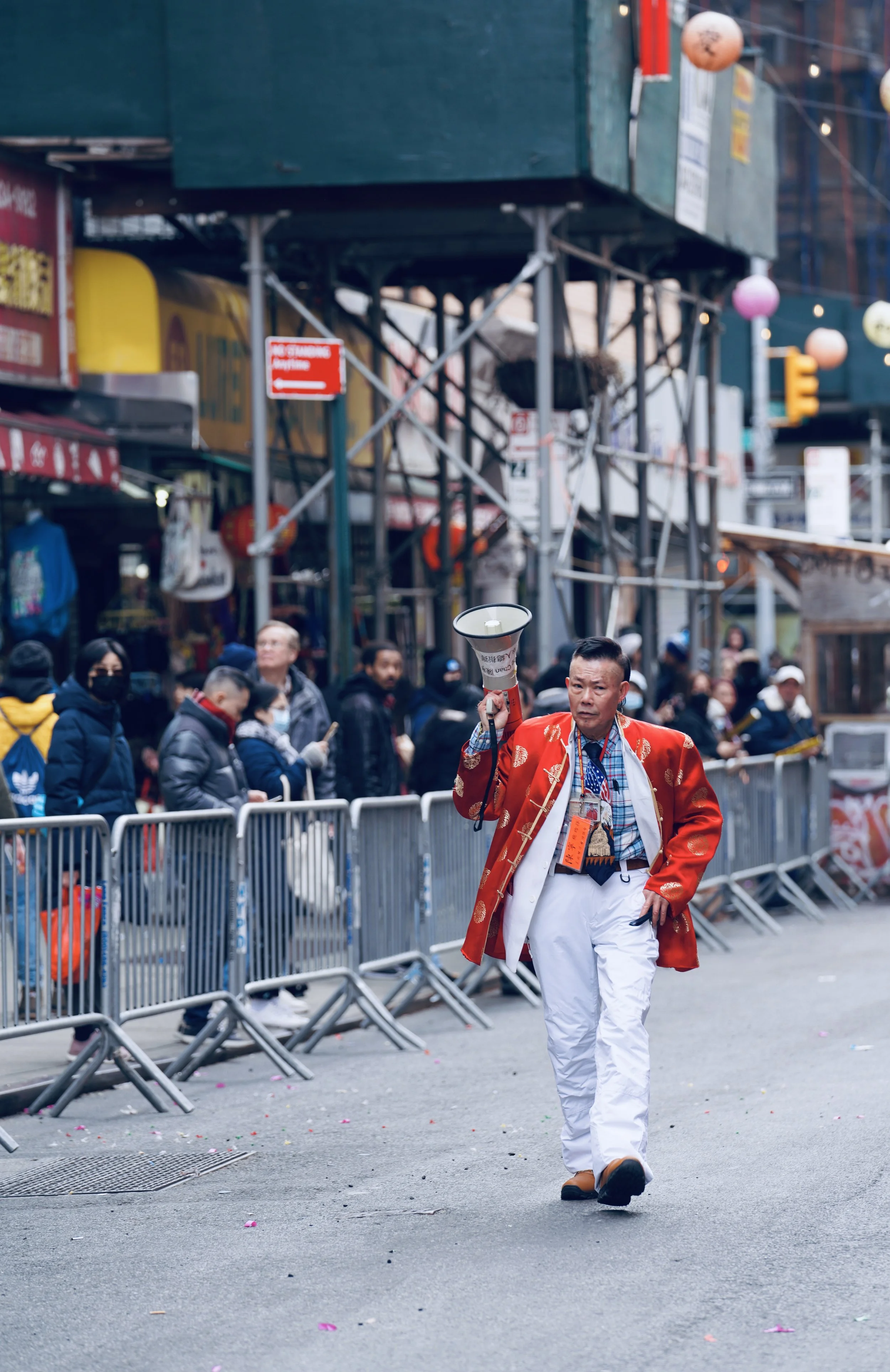 A man dressed in traditional Asian attire, holding a megaphone, walking past a crowd behind metal barricades on a city street.