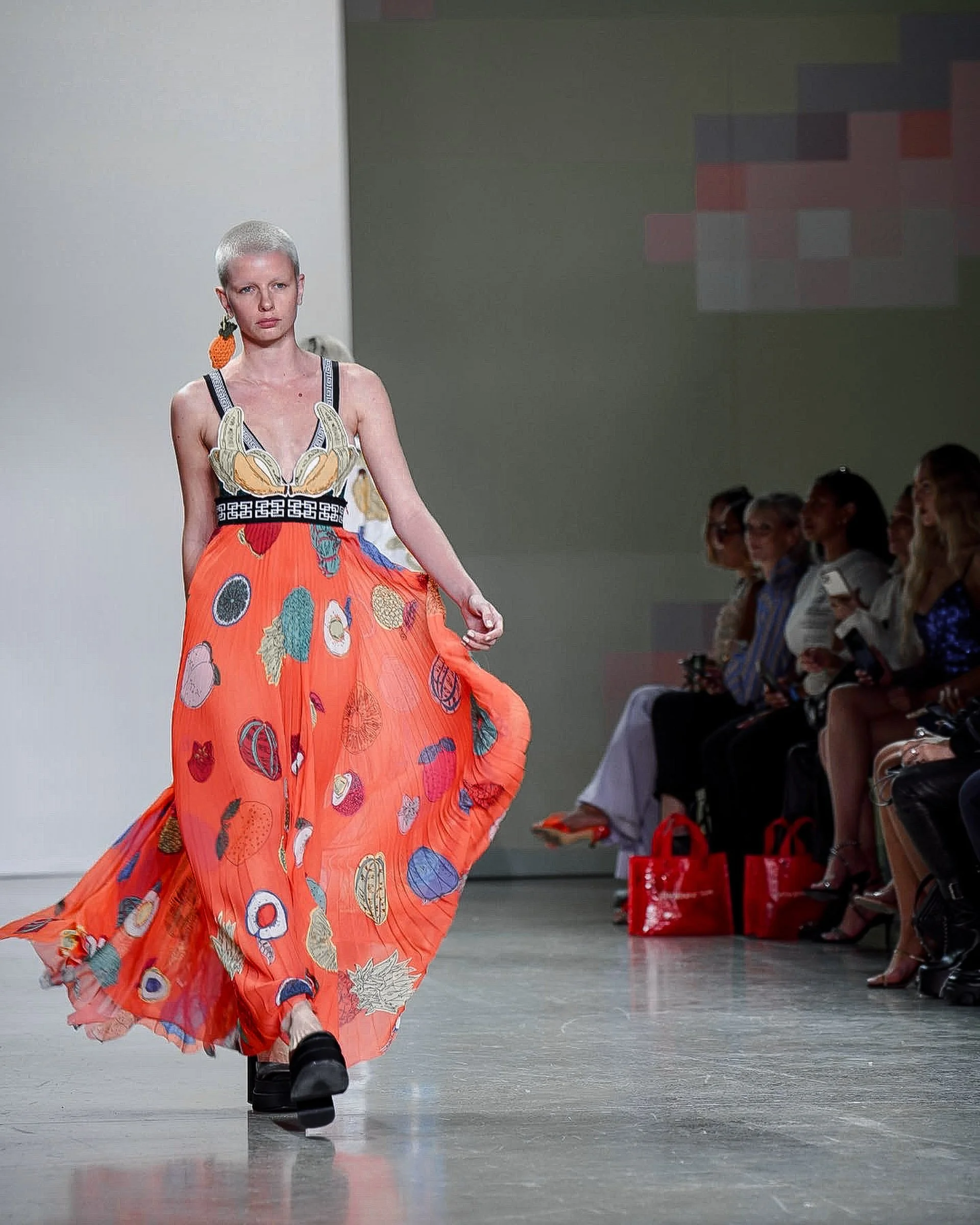 A model is walking down the runway wearing a colorful, patterned dress with a mixture of fruit and abstract designs, and chunky black shoes. The background shows a seated audience and a plain wall.