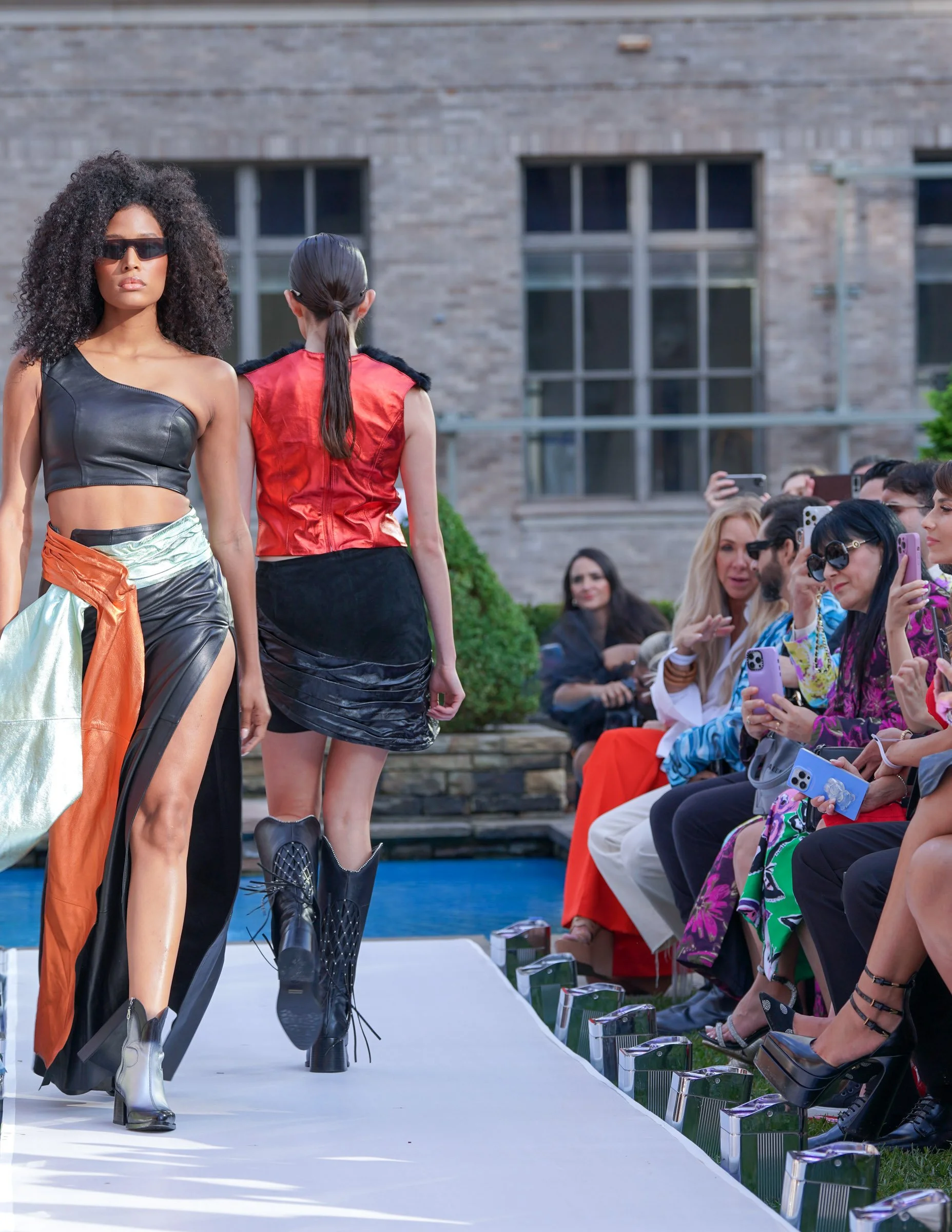 Models walking down a runway at a fashion show, with audience members seated on either side, taking pictures with smartphones.