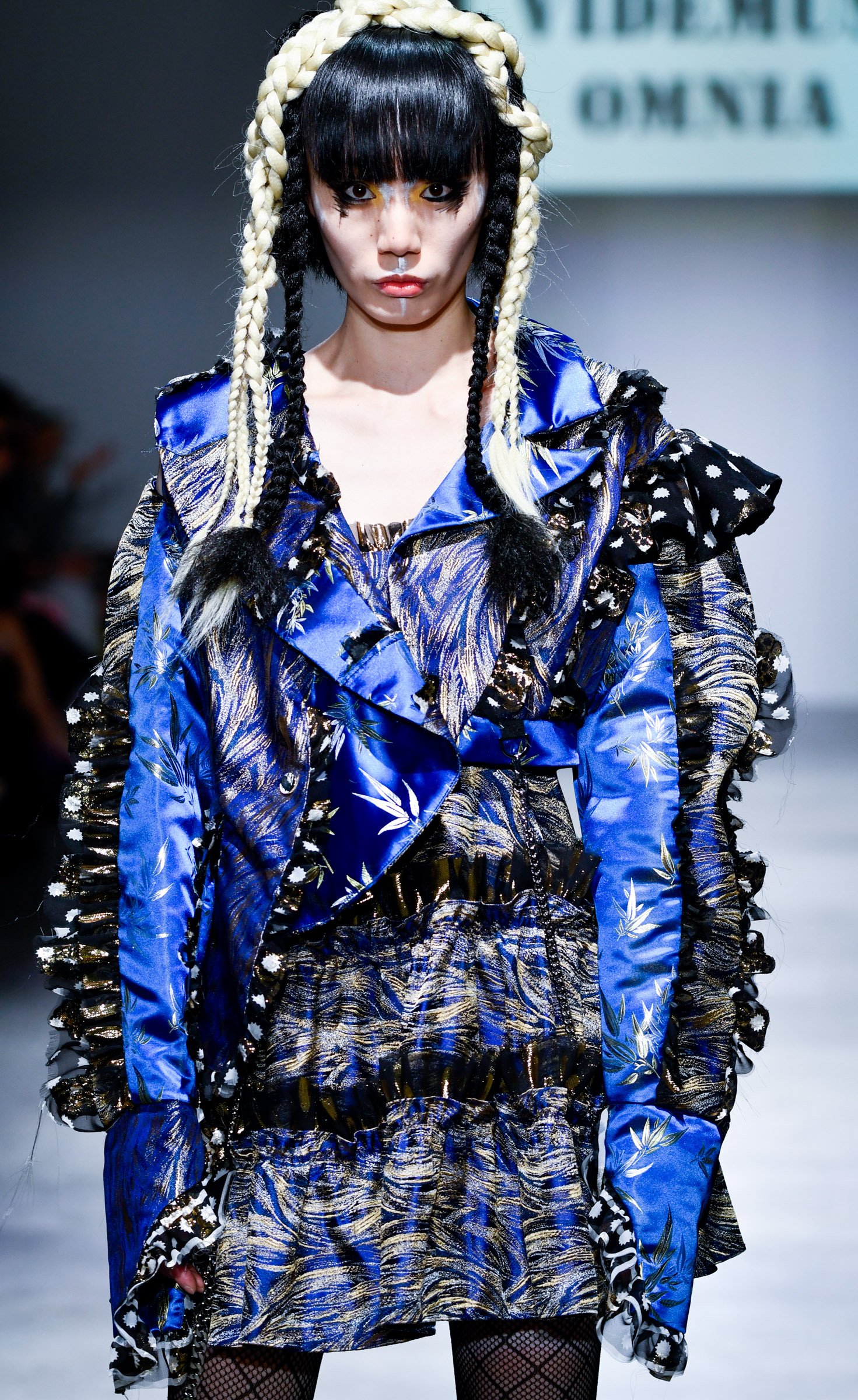 A female model walking on a runway wearing a blue and black patterned oversized jacket and a matching skirt with gold accents, fishnet stockings, and black gloves. She has long black hair with braided blonde extensions, bold makeup with yellow eye sh