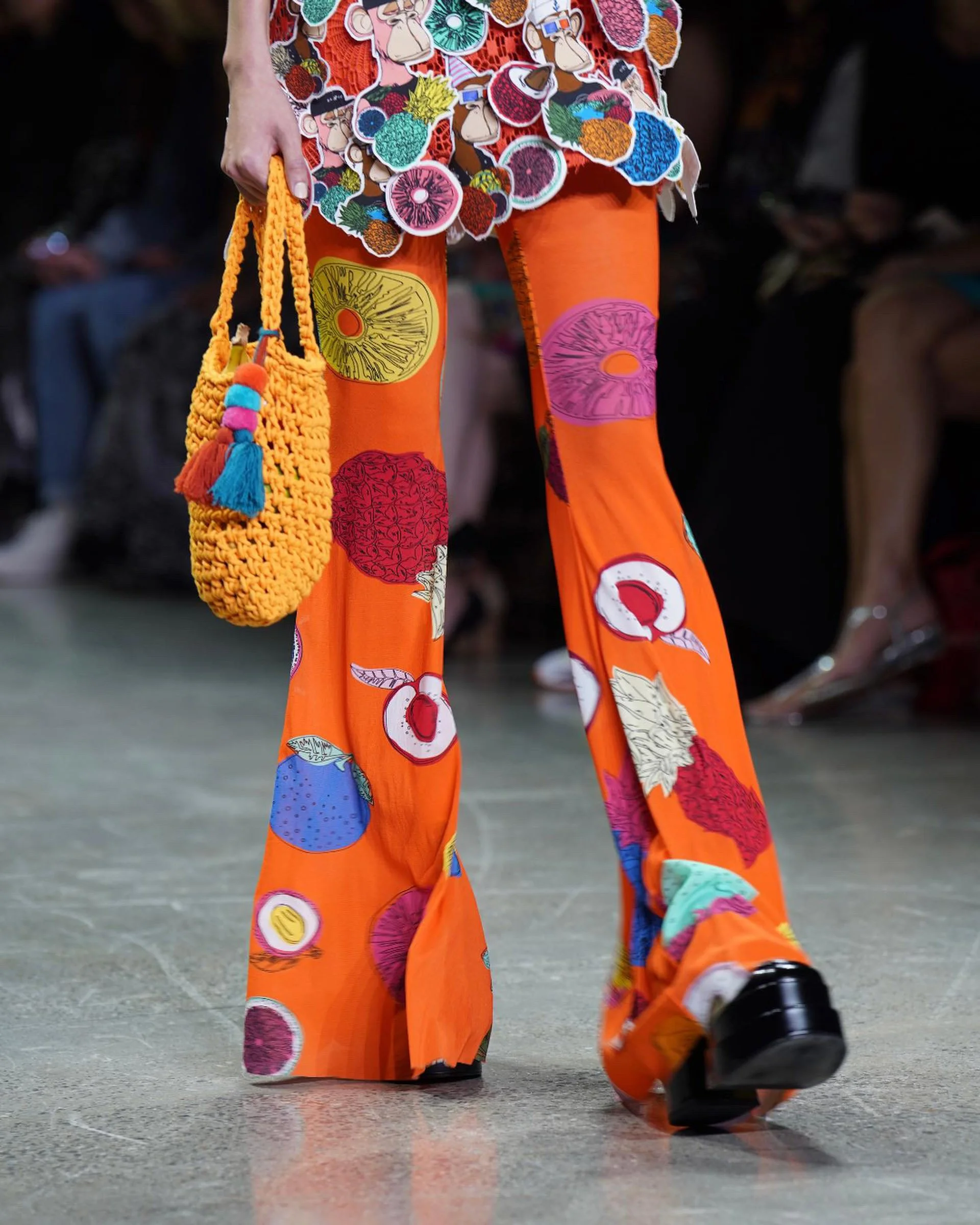 Close-up of a person wearing bright orange pants decorated with colorful fruit illustrations, walking on a floor with a textured surface, carrying a yellow crocheted bag with beads and a tassel.