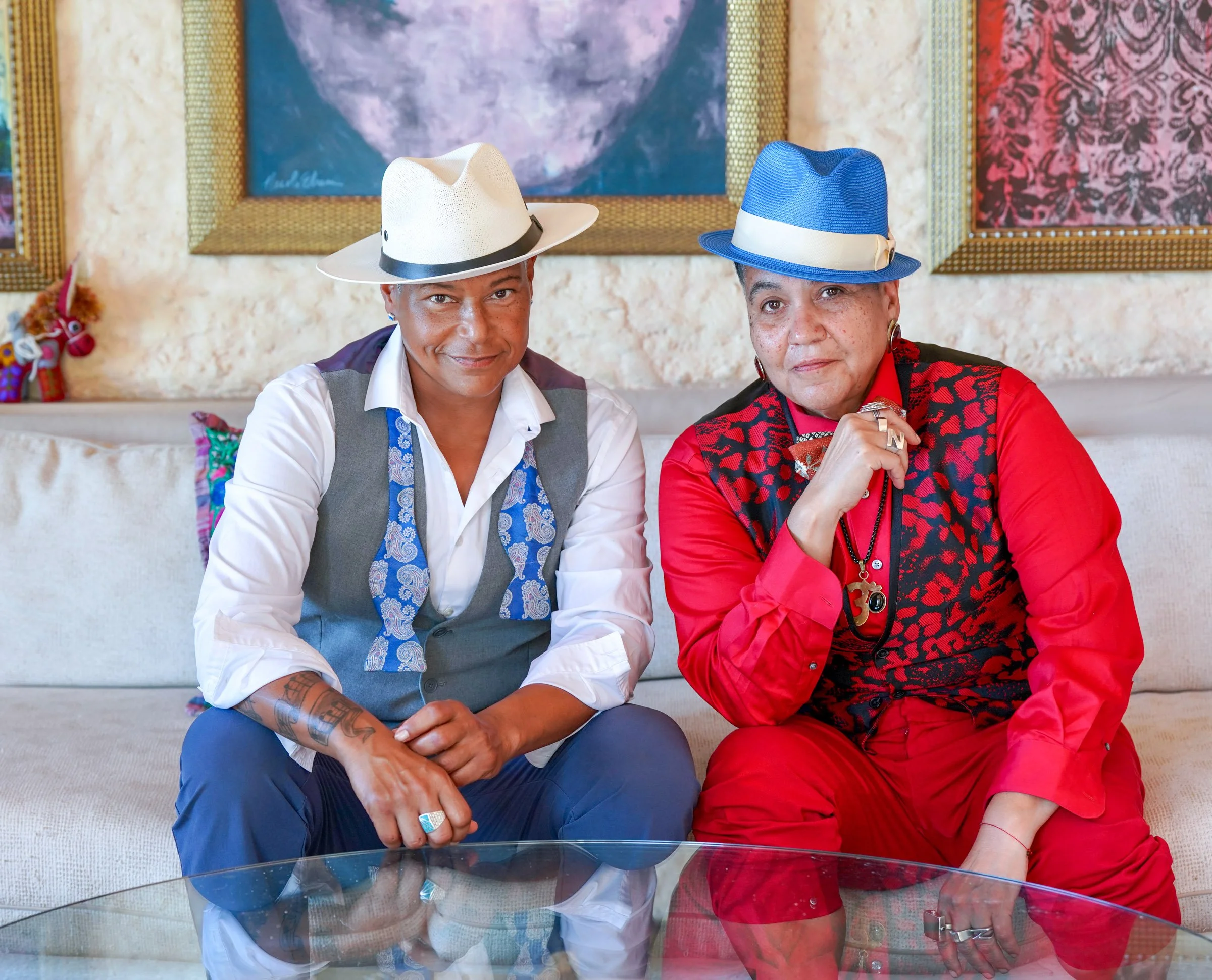 Two people, one wearing a white hat and a white shirt, the other wearing a blue hat and a red vest, sitting on a sofa with paintings on the wall behind them.