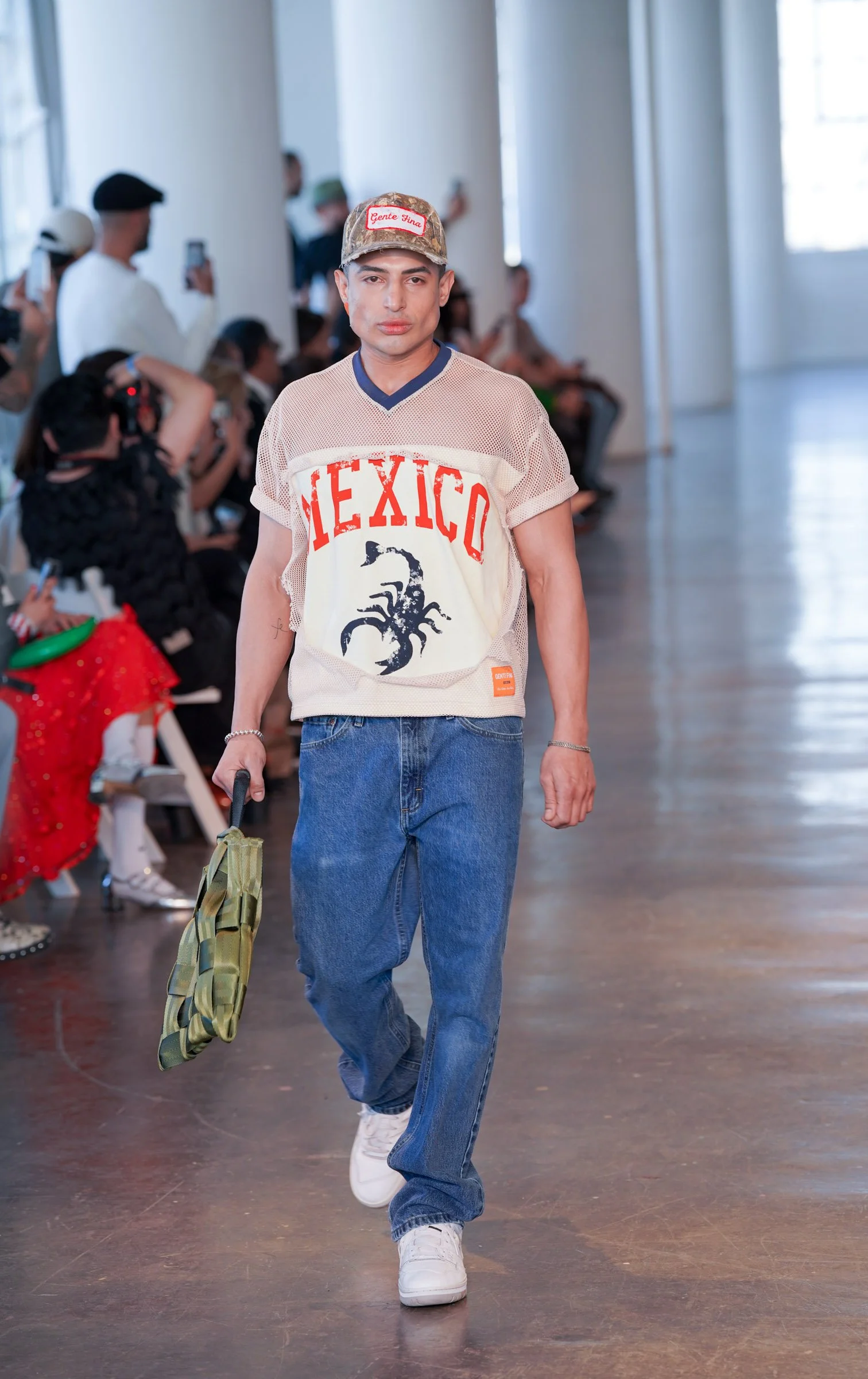 Man walking in indoor space, wearing a beige mesh t-shirt with 'Mexico' and a black scorpion graphic, blue jeans, white sneakers, pink cap with red patch, holding a green camouflage bag.
