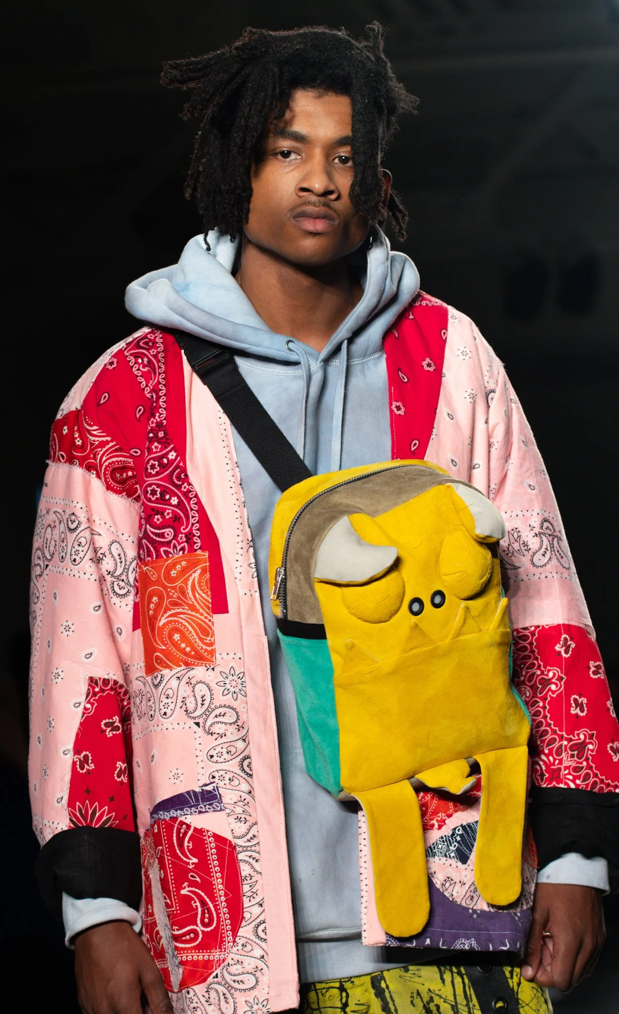 A young man with dreadlocks wearing a colorful jacket and hoodie, carrying a yellow plush backpack, standing against a dark background.
