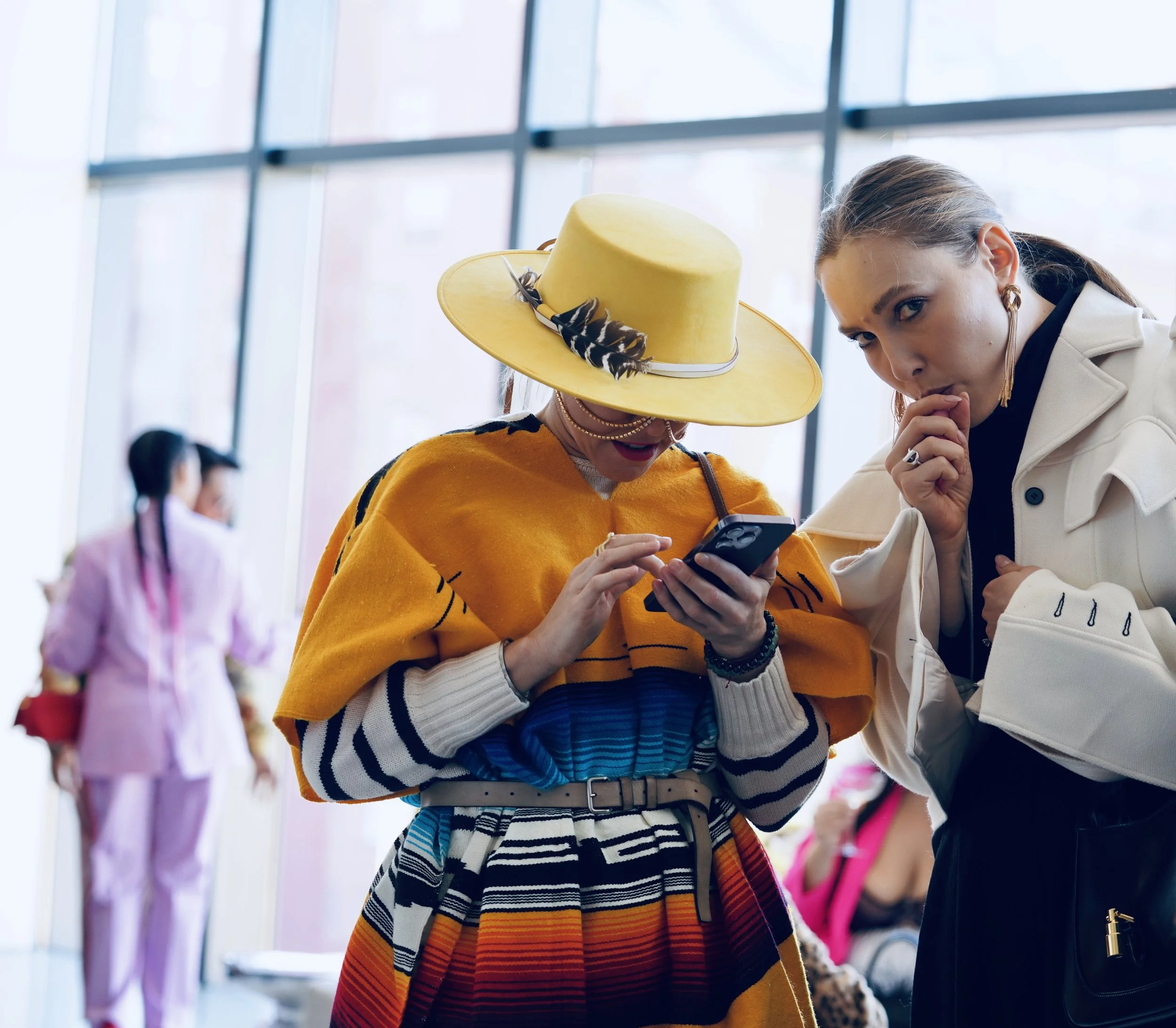 Two women are closely examining a smartphone together, with one wearing a yellow hat decorated with feathers and a yellow top, and the other wearing a white jacket and large hoop earrings, in a bright, modern indoor space with large windows.