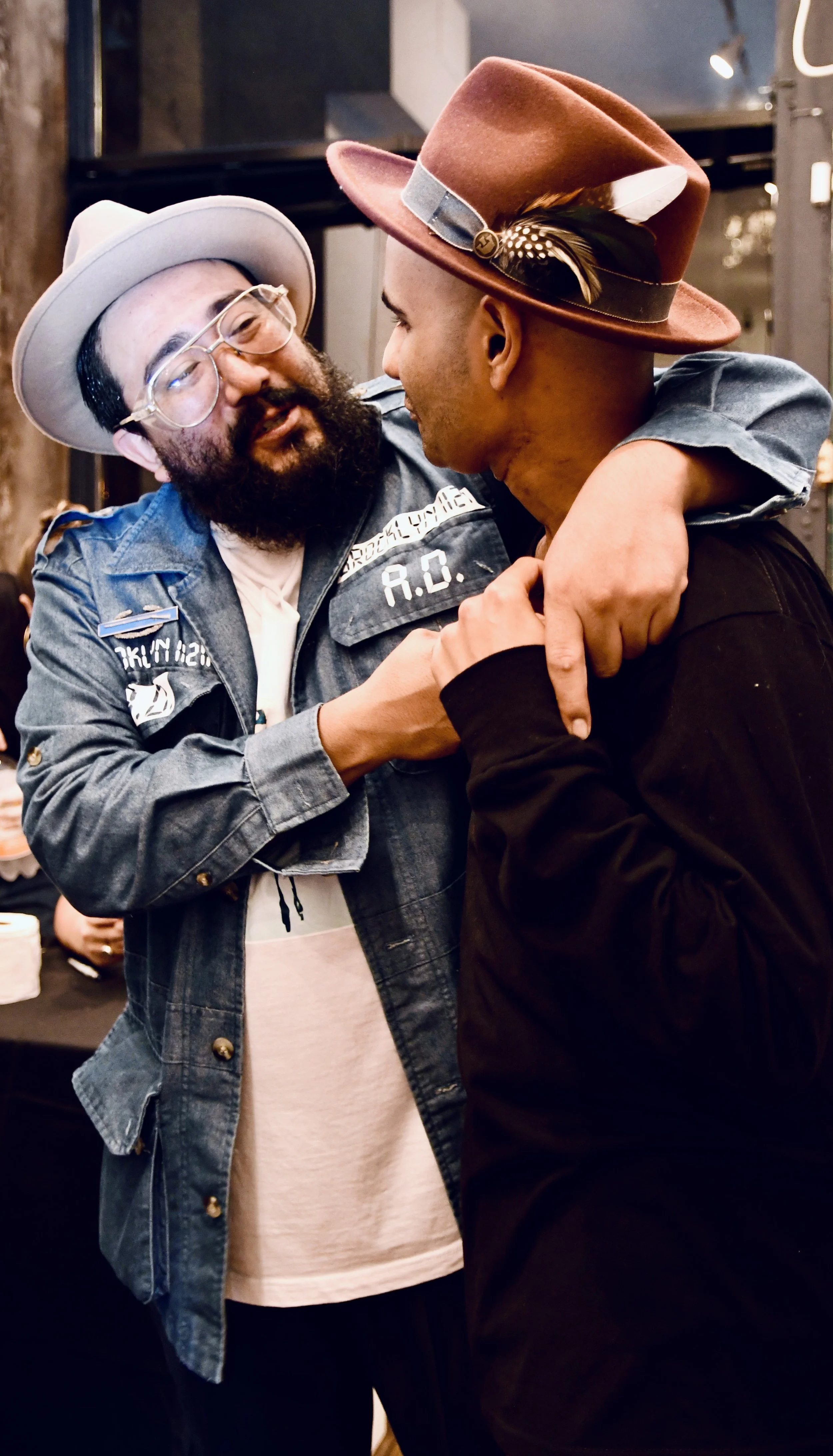 Two men with hats and facial hair joyfully interact indoors, with one man wrapping his arm around the other's shoulder.
