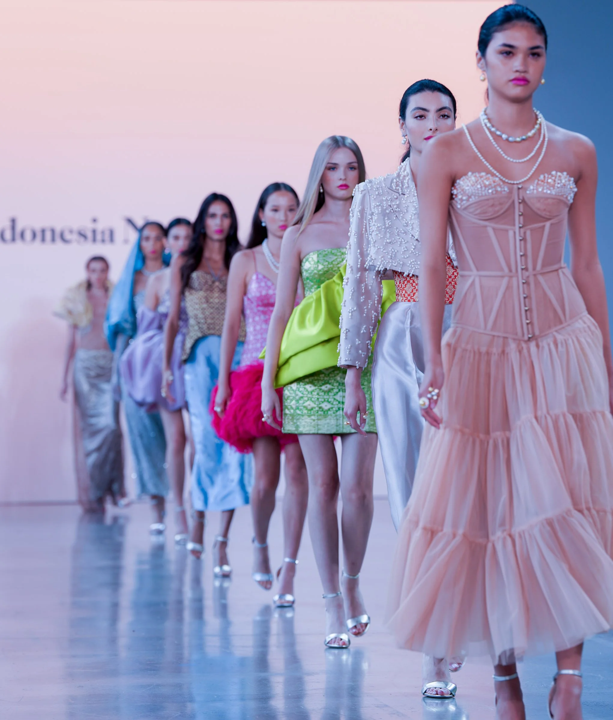 A line of models walking on a runway during a fashion show, dressed in elegant, colorful dresses with jewelry and high heels.