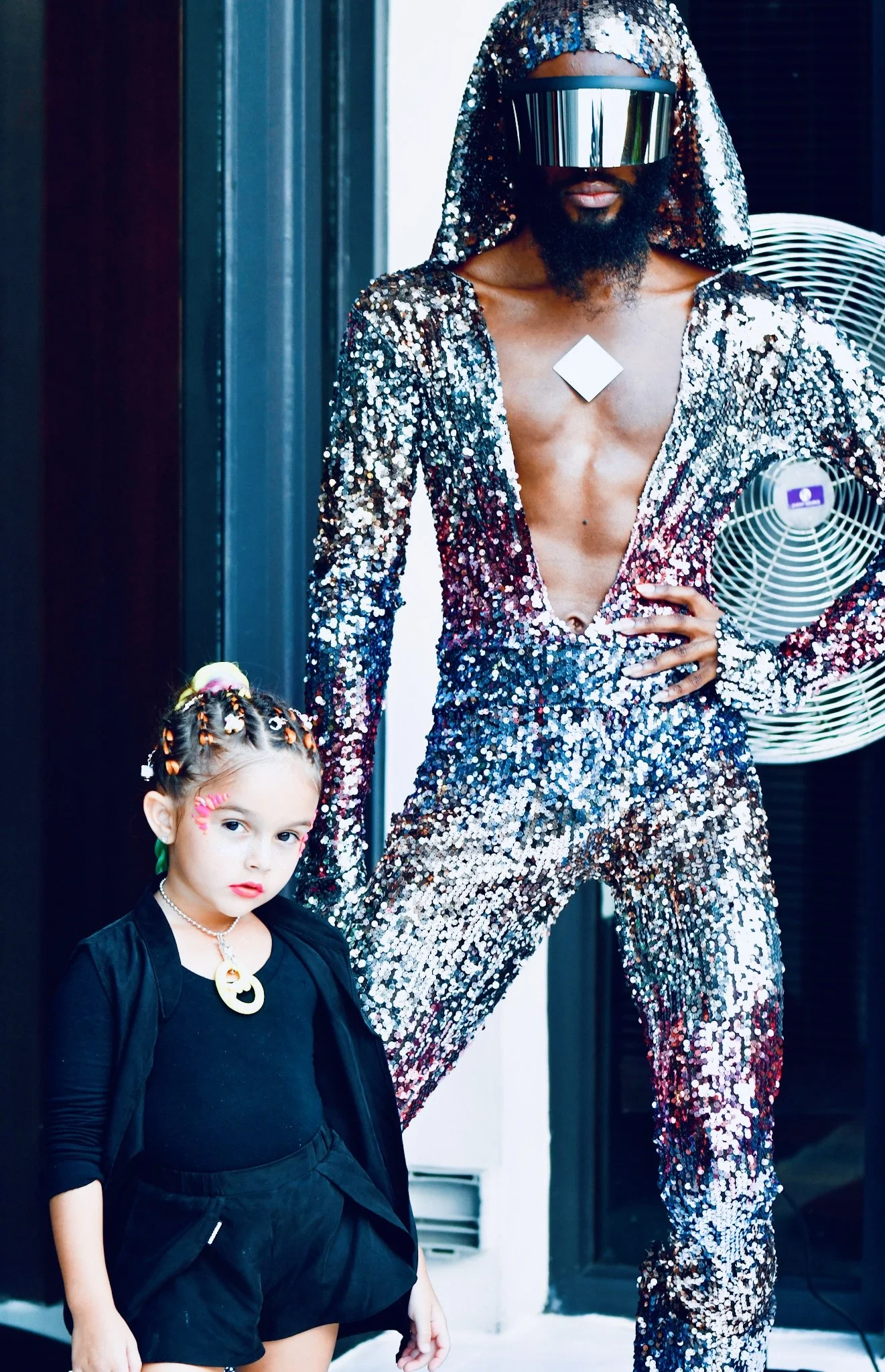 A man wearing a sequined, deep V-neck suit and a matching hooded jacket, with a reflective visor covering his eyes, standing with a hand on his hip. Next to him is a young girl with braided hair decorated with beads, wearing a black outfit with a Bat