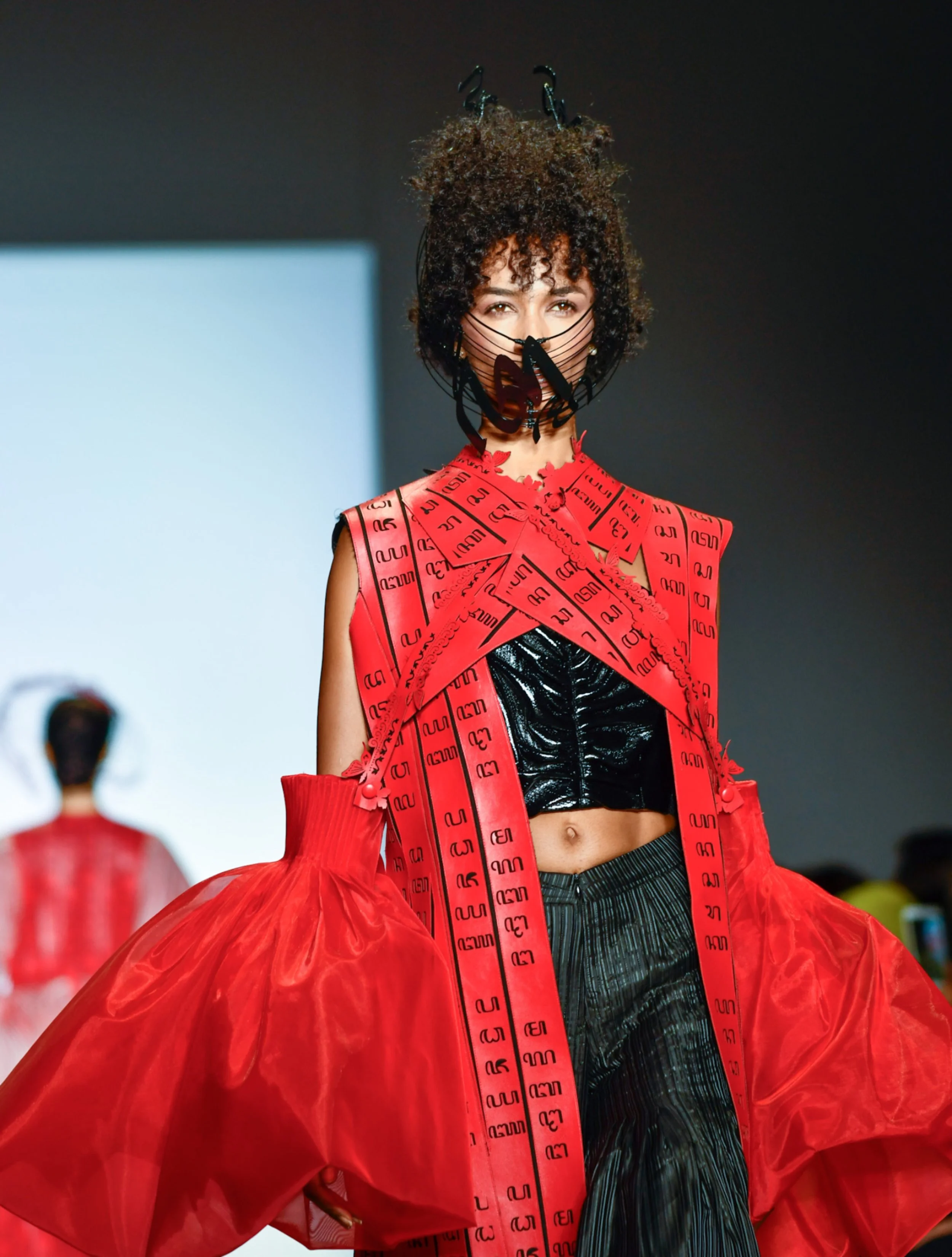 Model wearing a red dress made from material resembling measuring tape, with a black shiny skirt underneath, on a runway. The dress has a voluminous red tulle layer. The model wears a black face mask with a butterfly design and has curly hair.