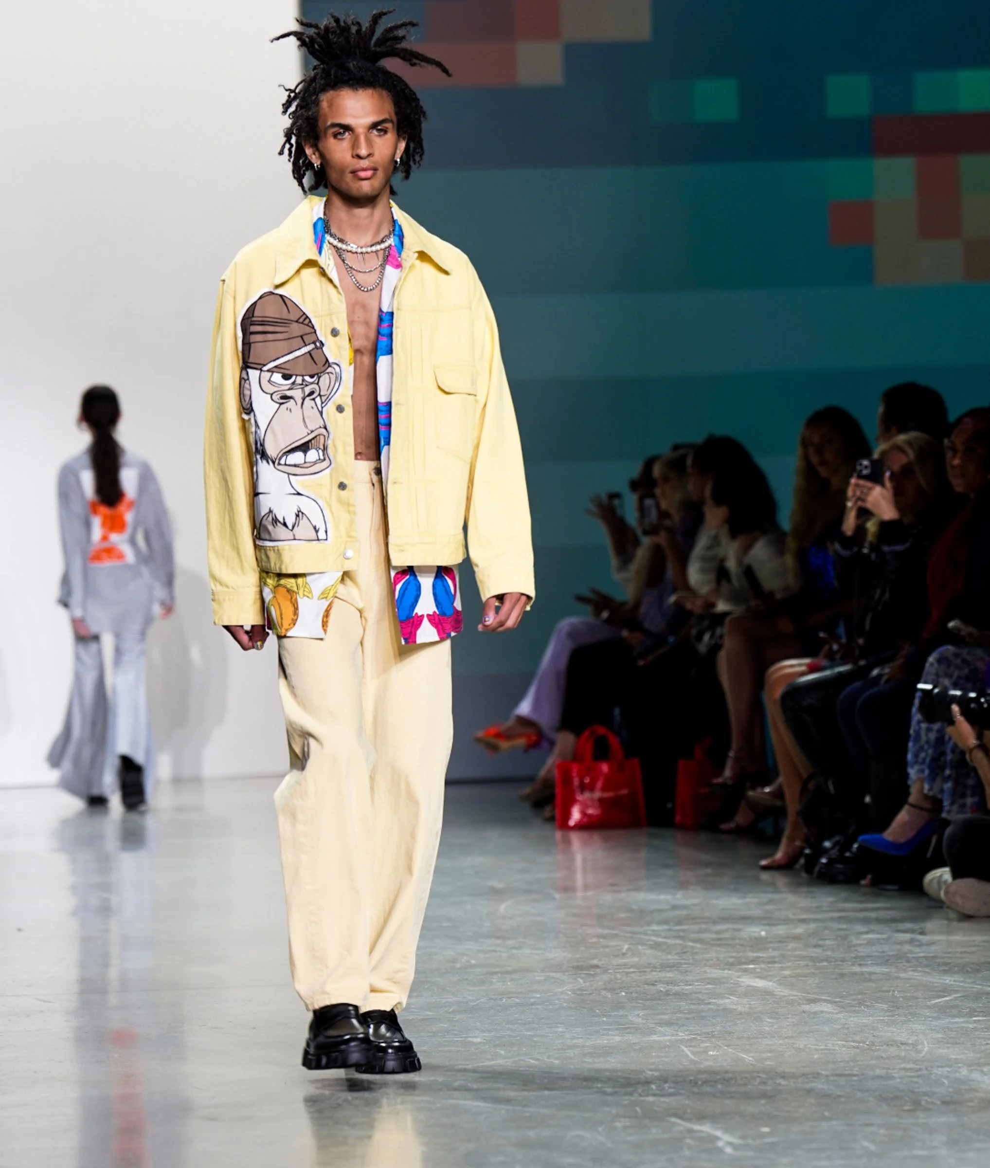 A male model walking on a runway during a fashion show, wearing a yellow jacket with a cartoon monkey printed on it, beige pants, and black chunky shoes, with audience members watching in the background.