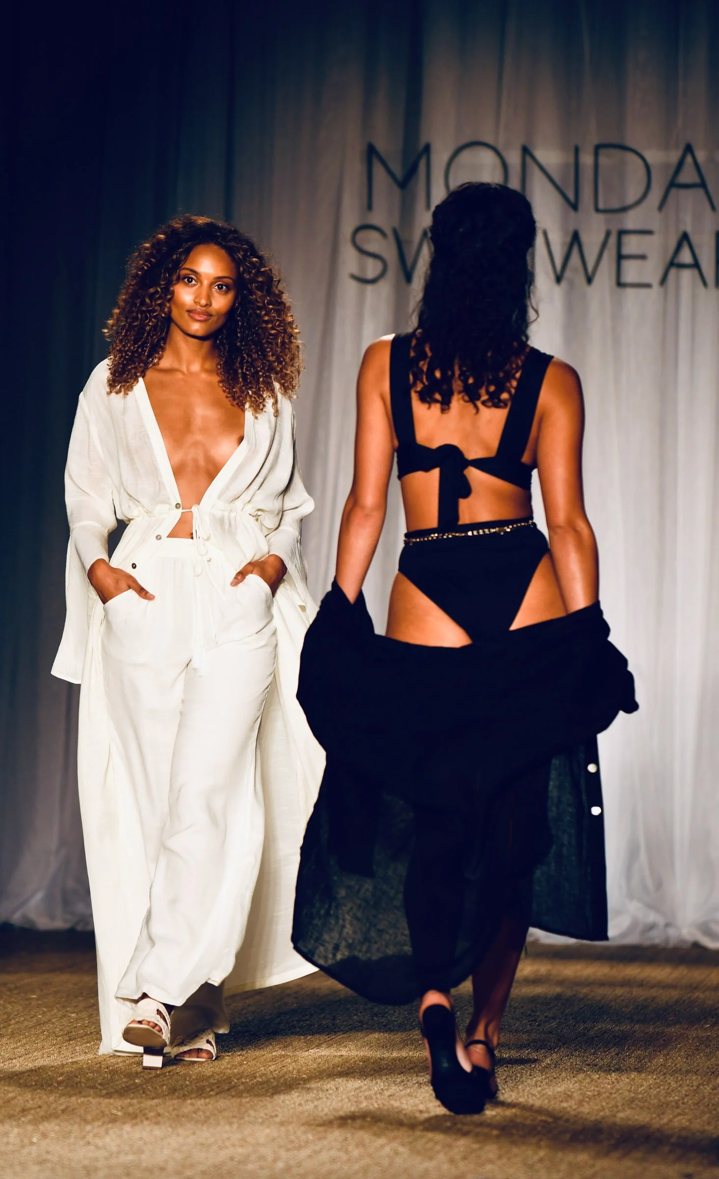 Two women walking on a runway during a fashion show, with one in a white outfit and the other in a black lingerie ensemble, against a backdrop with the text 'MONDAY SWIMWEAR'.