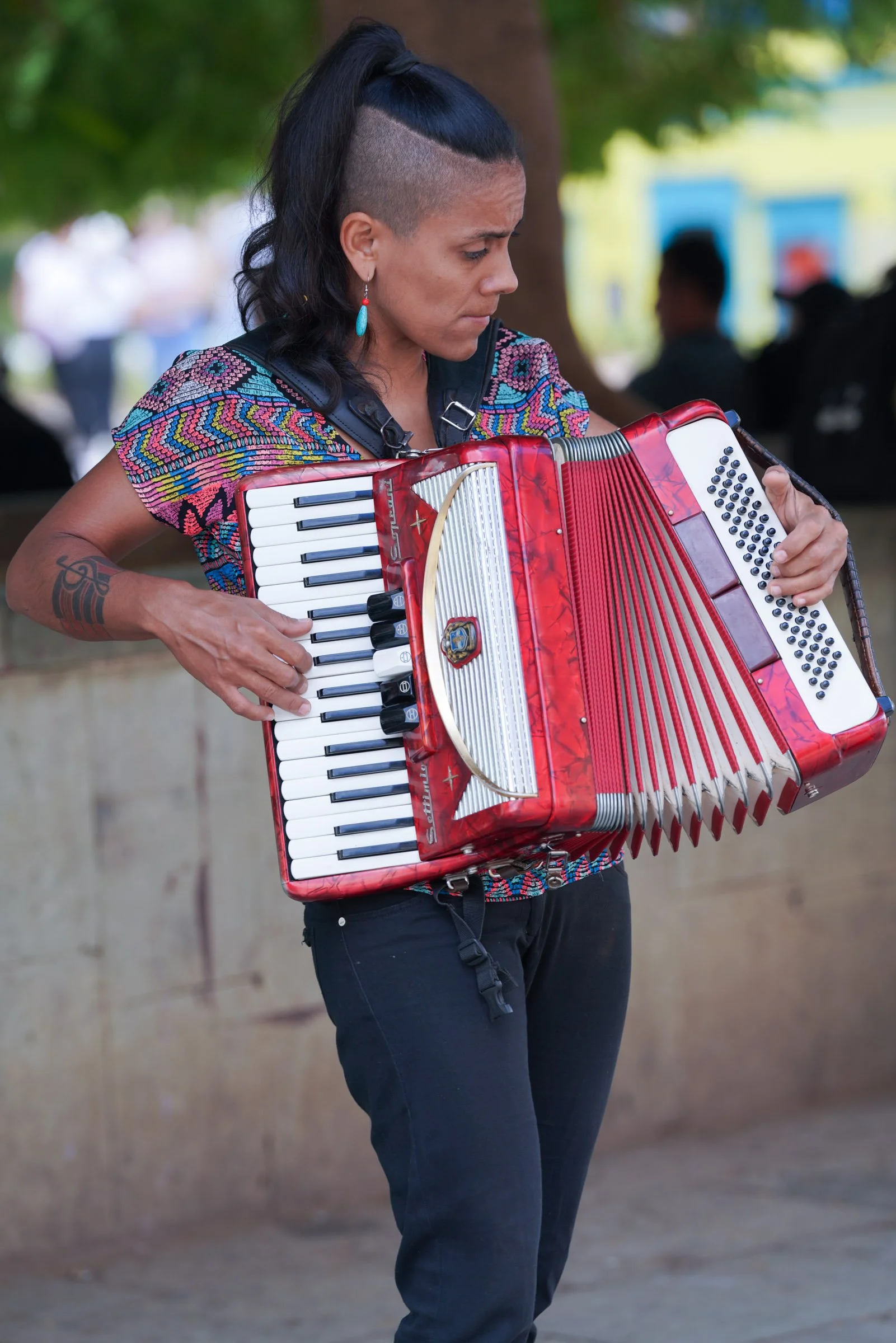 A woman with dark hair styled with a shaved side and a long ponytail on top, playing a red and white accordion outdoors near a tree, wearing a multicolored embroidered shirt and earring with a turquoise stone.