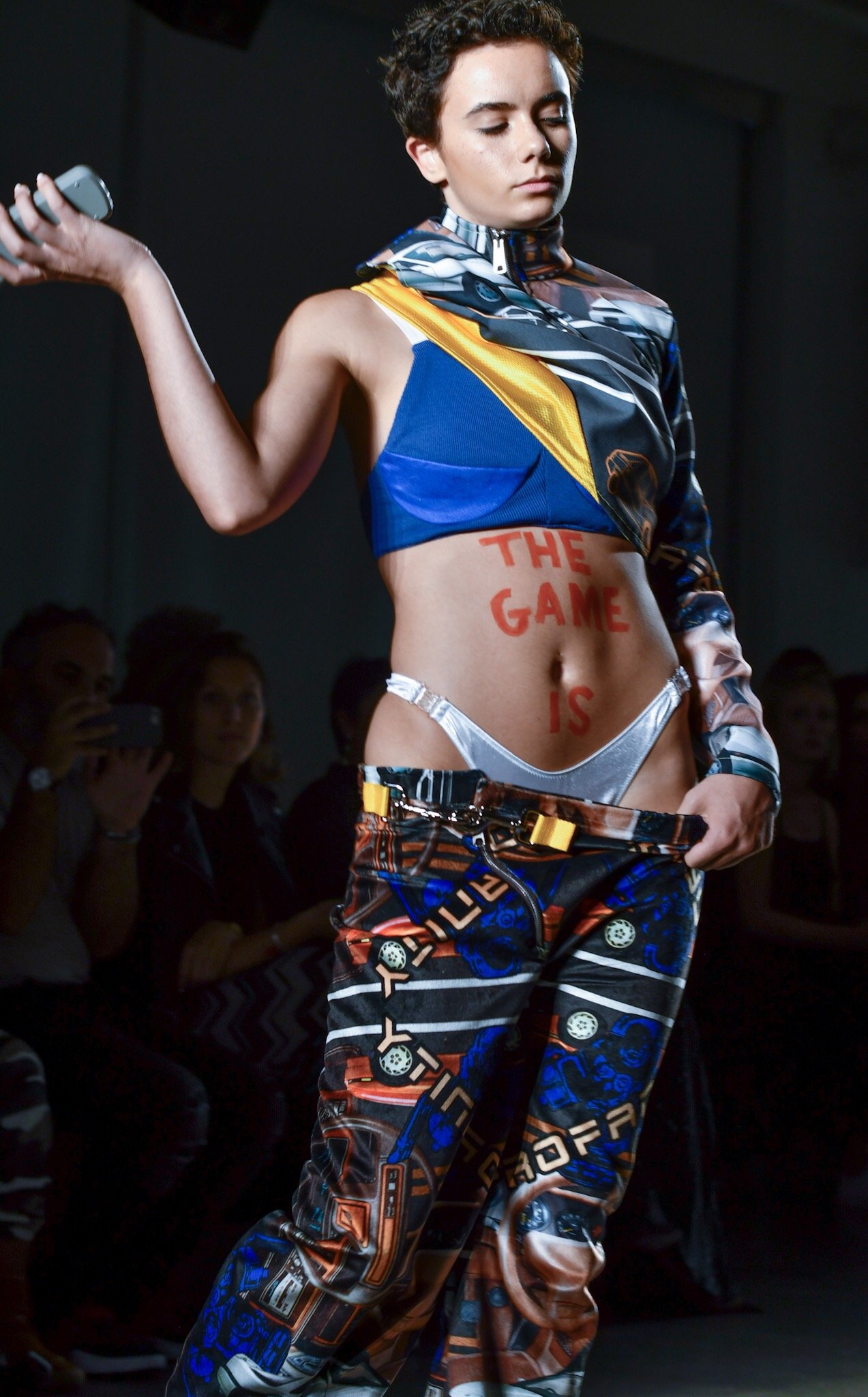 A young woman with short, curly hair and freckles models on a runway. She is wearing a colorful, patterned outfit with a jacket and matching pants. The phrase 'THE GAME IS' is written in red on her stomach. She has a confident pose, with one arm bent