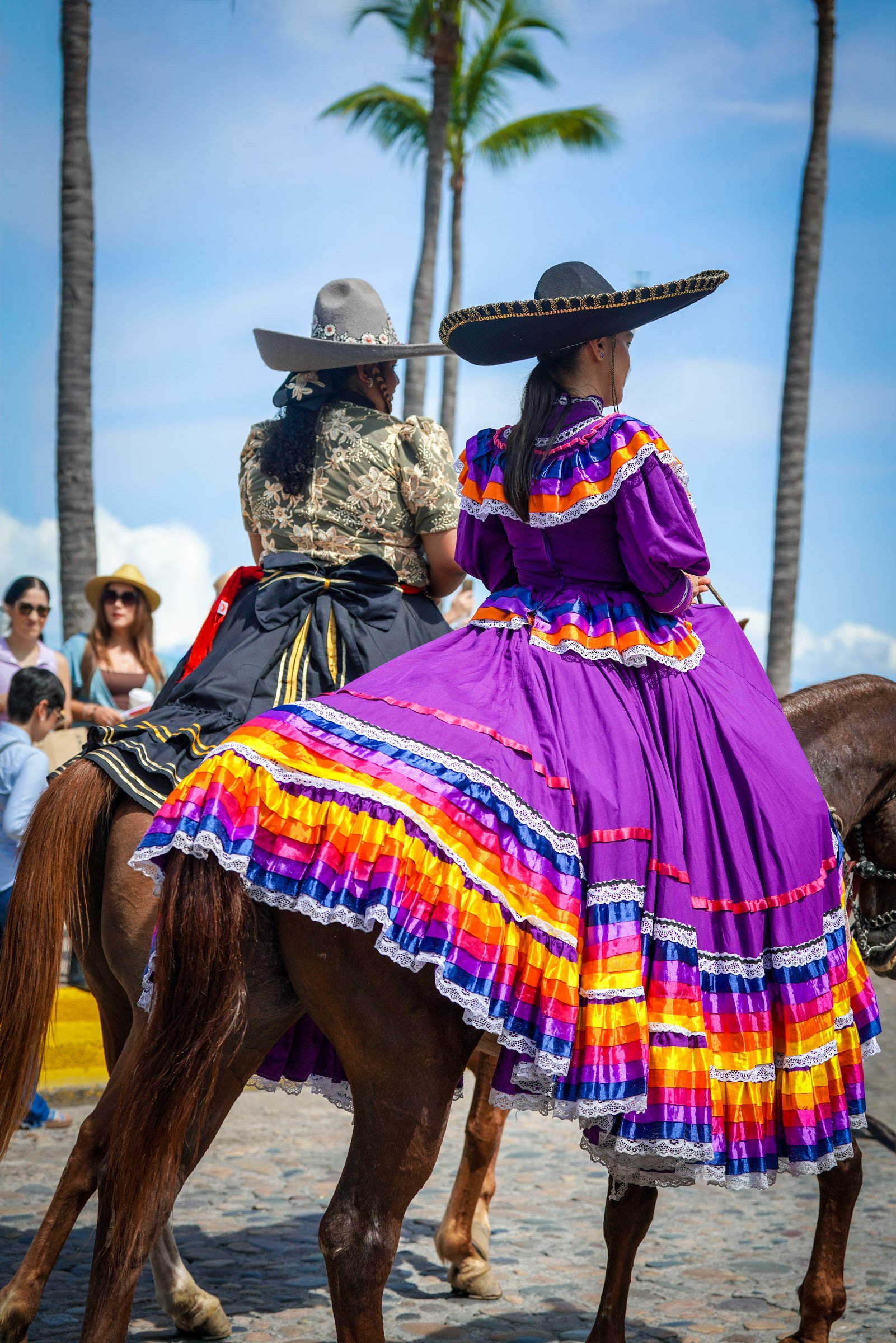 Two women wearing traditional Mexican dresses and large sombrero hats, riding horses during a parade, with palm trees and a crowd in the background.