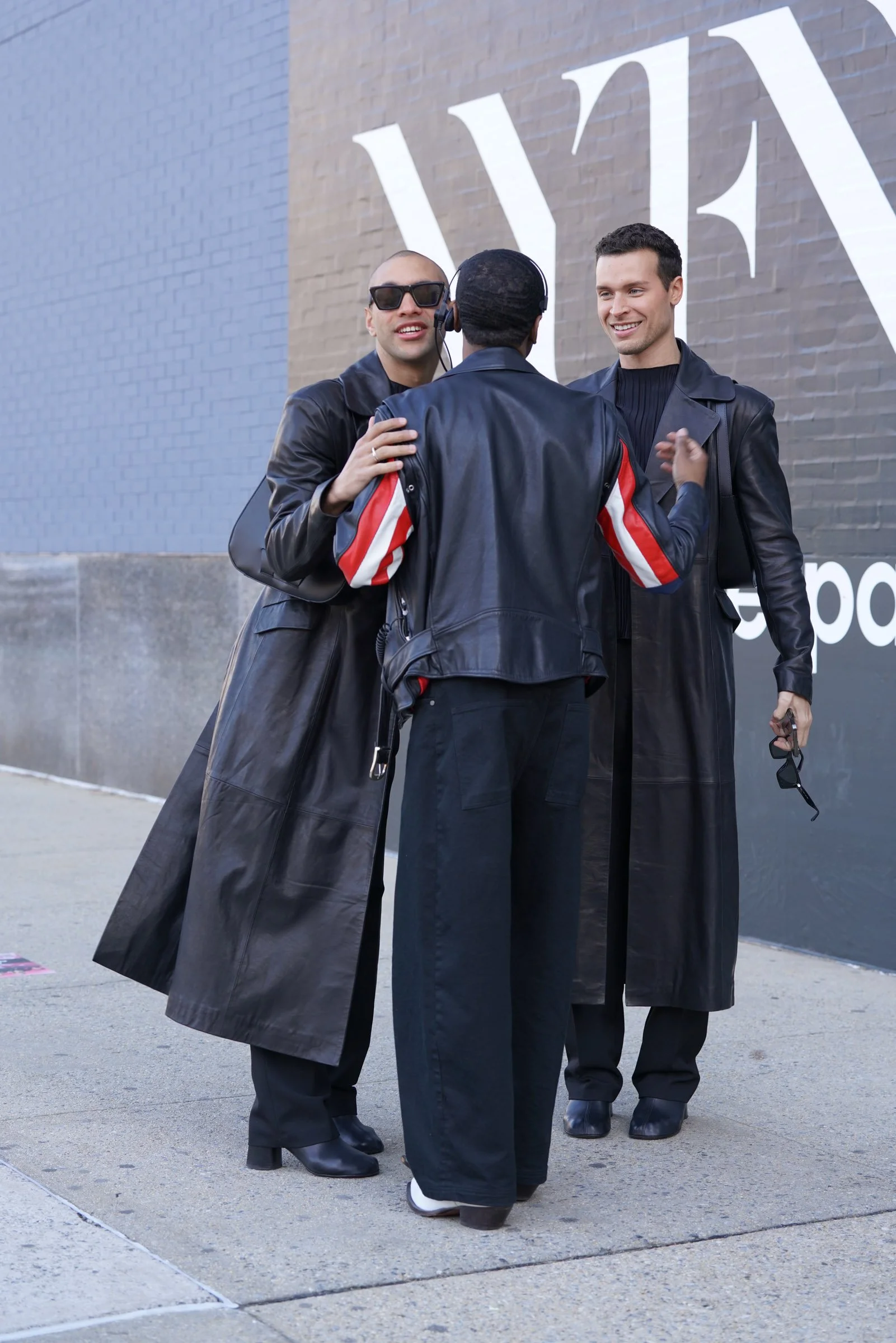 Three people standing outdoors in front of a wall with large white text, engaging in conversation with one person wearing a headset, all dressed in black leather coats and black boots.