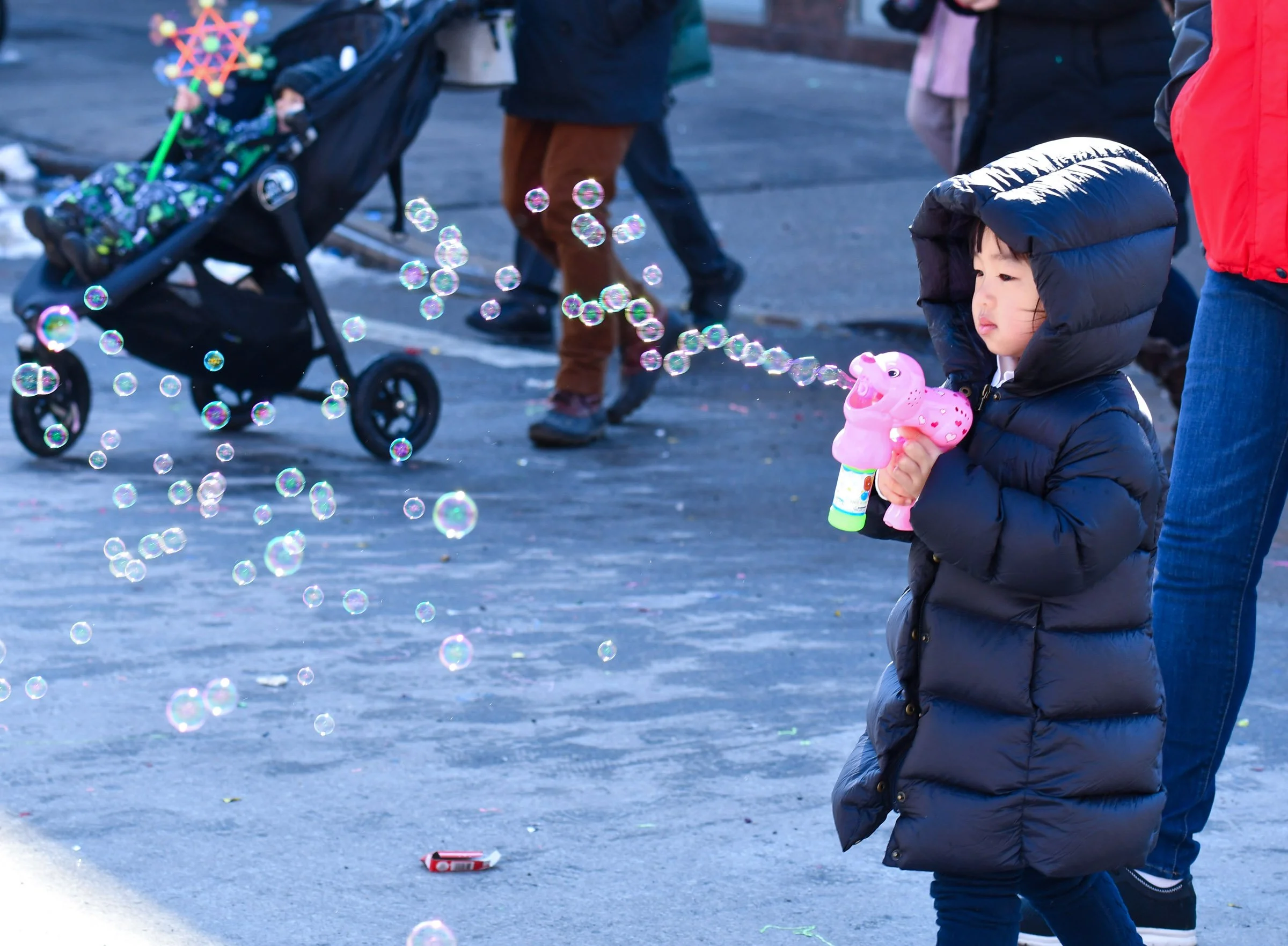 A young child dressed in a black winter coat with a hood, holding a pink toy that looks like a hippopotamus, standing on an outdoor pavement. There are bubbles floating in the air around the child, and other people walking nearby, some with strollers