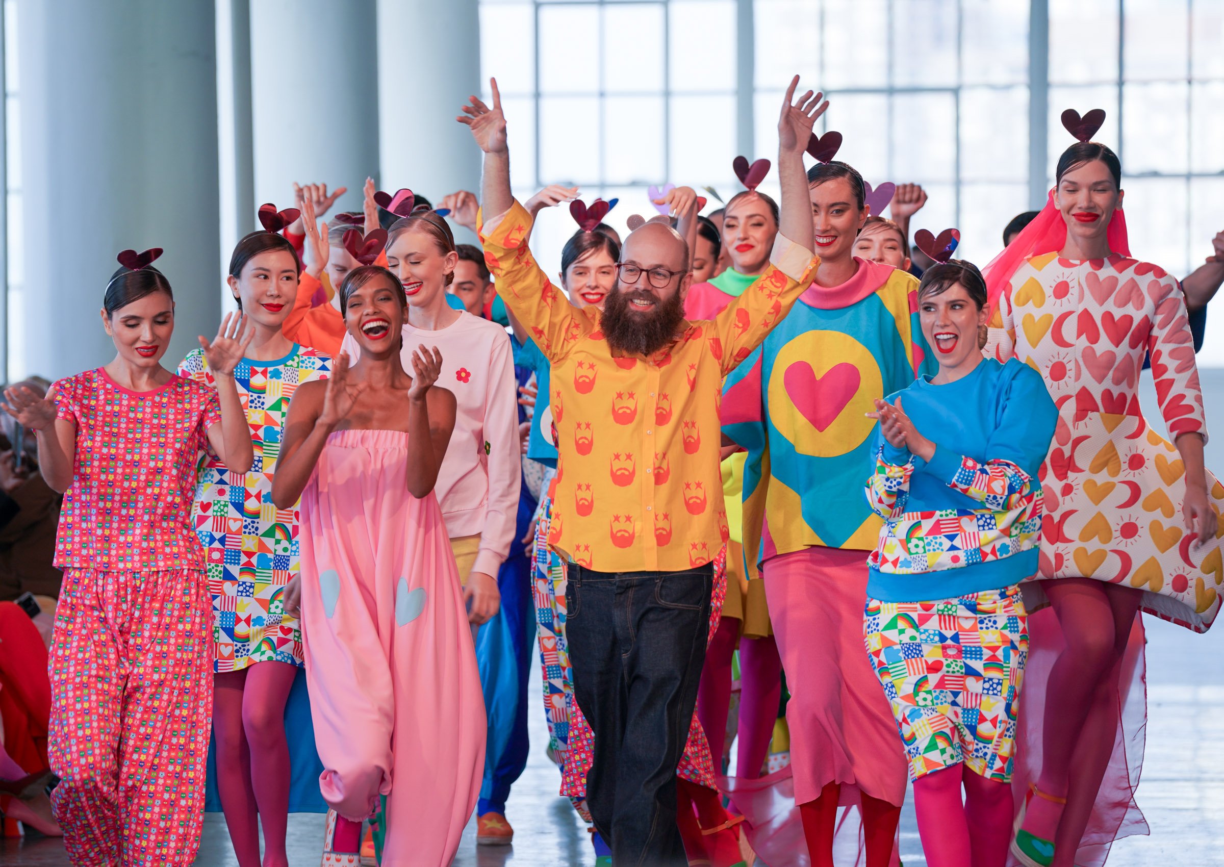 A diverse group of people dressed in colorful, playful costumes with heart and love-themed patterns, celebrating together indoors with large windows and bright natural light.