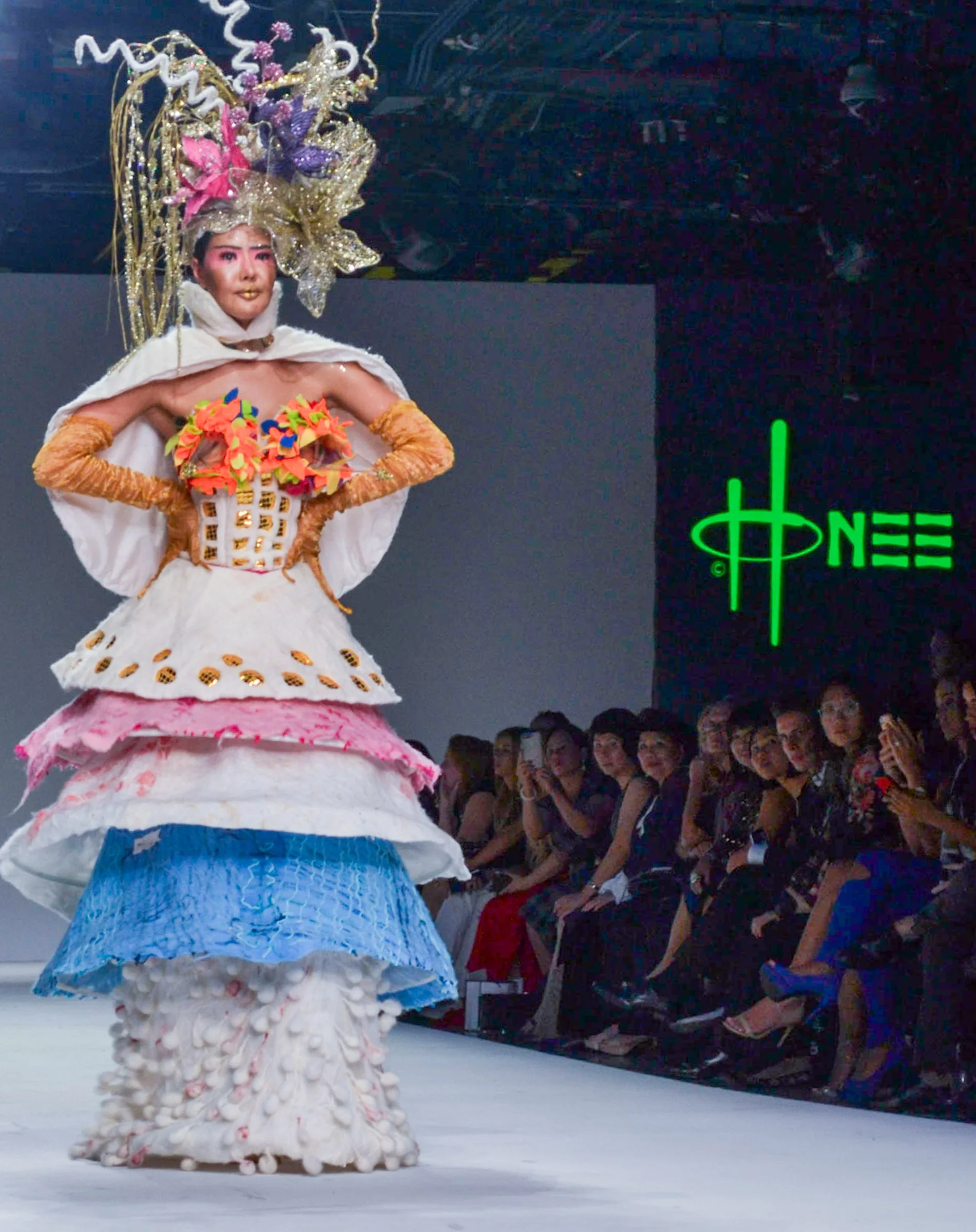 A performer in an elaborate costume with a tall, intricate headpiece and a dress adorned with colorful decorations and textured layers, standing on a stage at a fashion or art show, with an audience seated and watching.