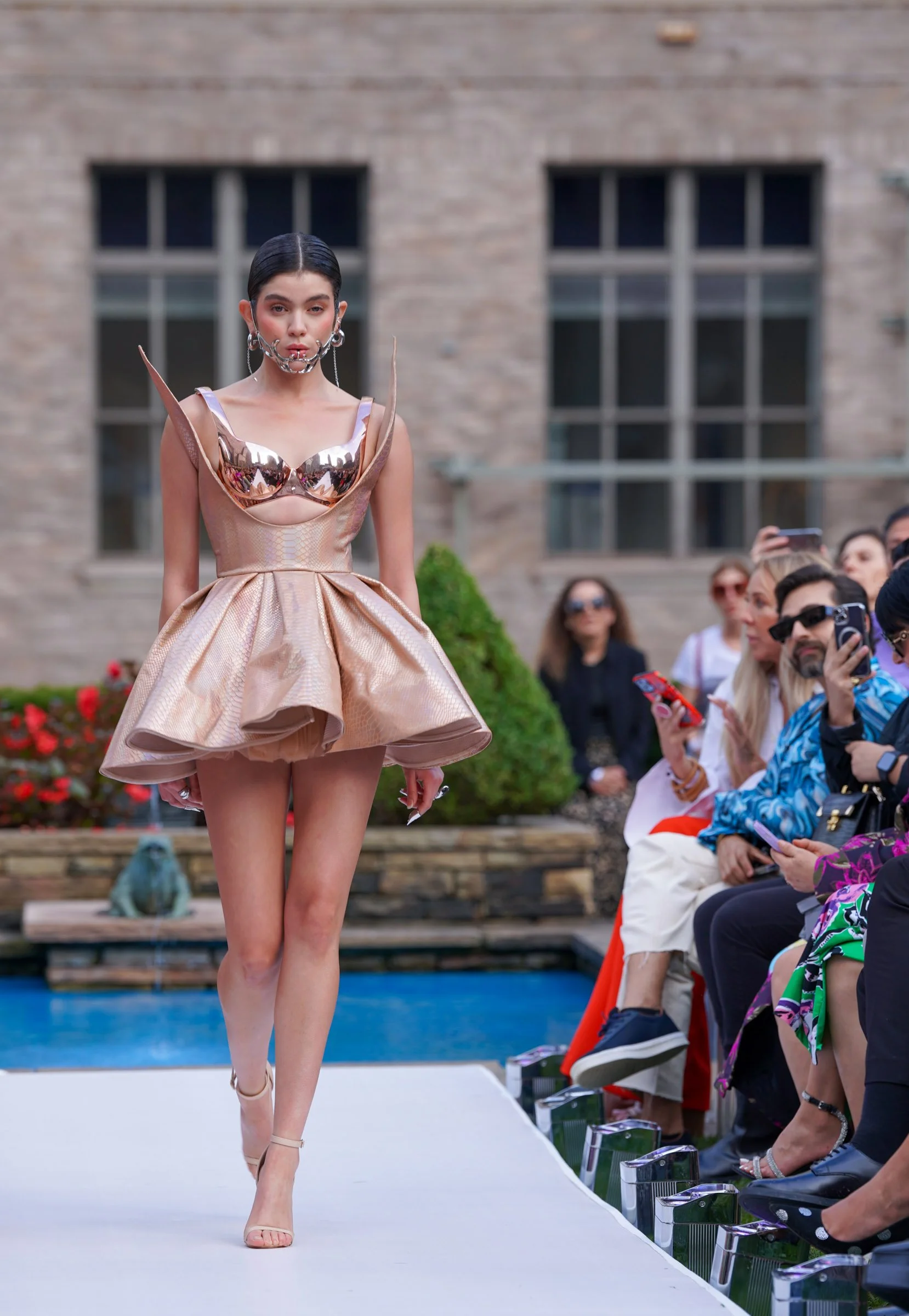 A female model walking on an outdoor runway during a fashion show, wearing a metallic, futuristic dress and high heels, with an audience watching.