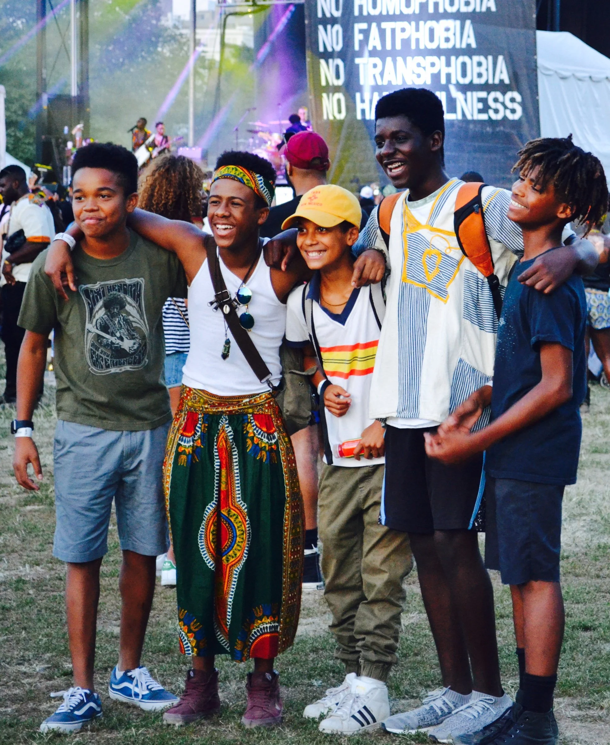 A group of five diverse young people standing close together at an outdoor event, smiling and with arms around each other's shoulders. In the background, a stage with musicians performing and a large screen displaying the words 'NO HOMOPHOBIA, NO FAT