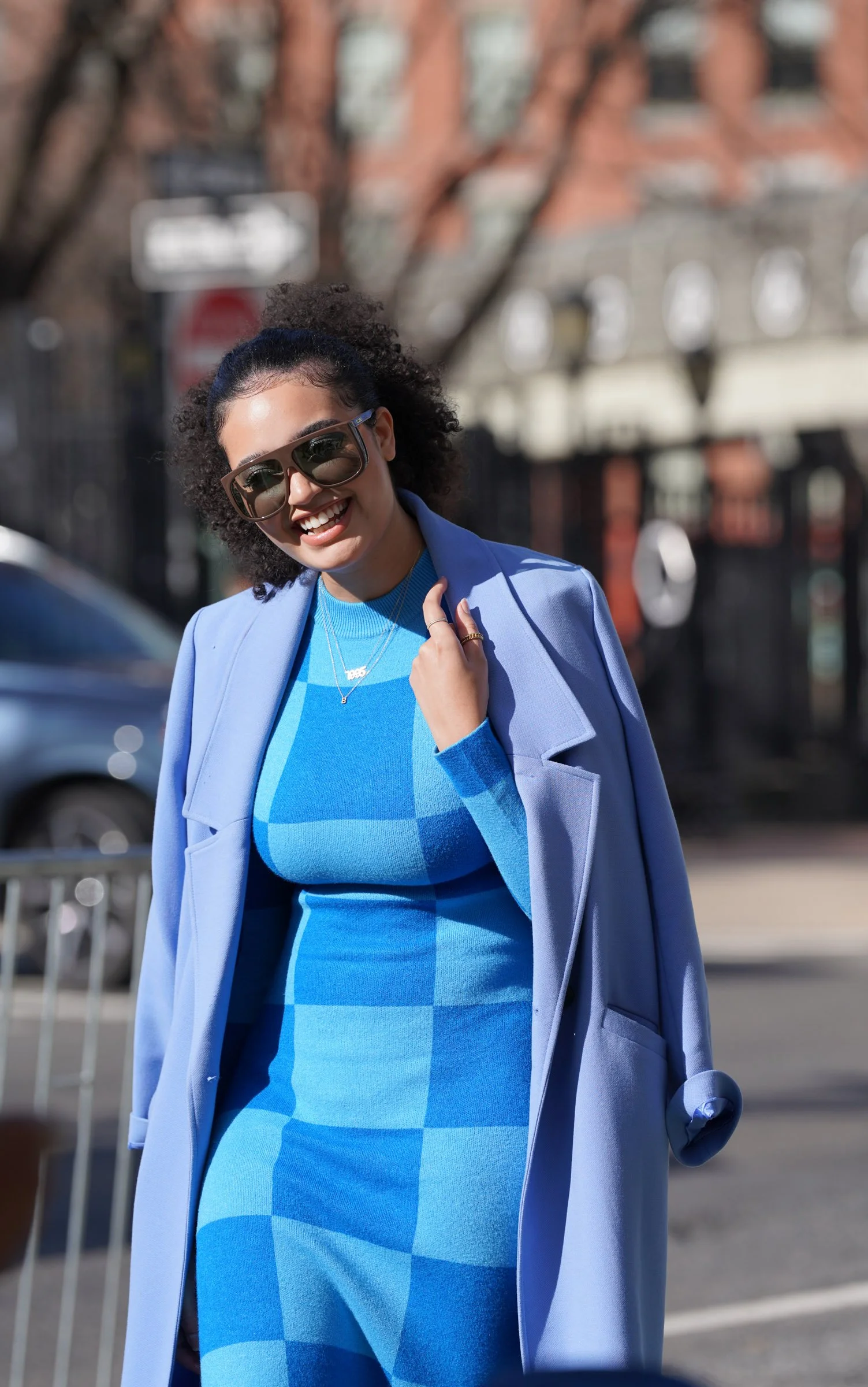 Woman with curly hair wearing sunglasses, a blue checkered dress, and a light blue coat, smiling outdoors.