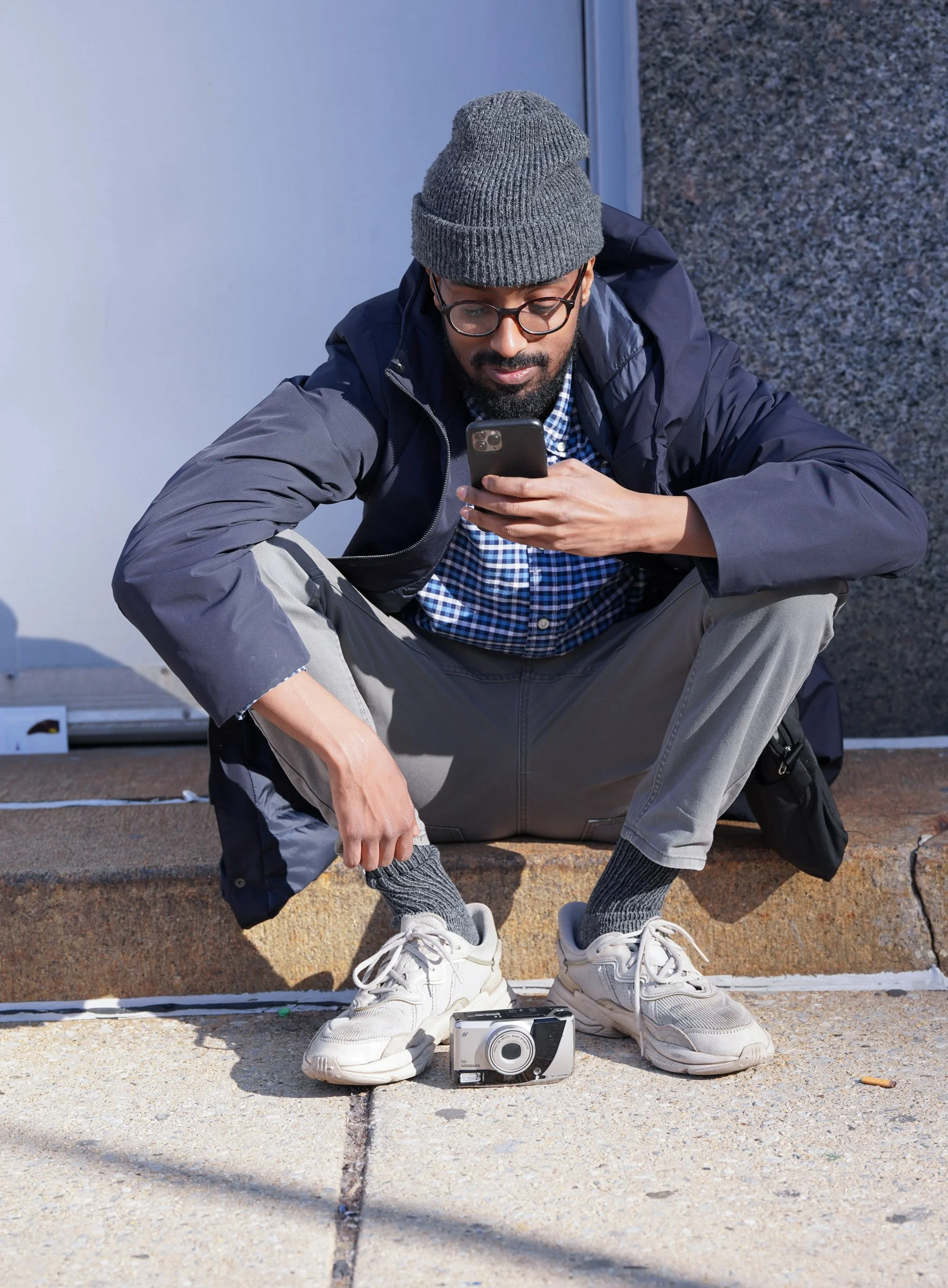 A man with a beard, glasses, gray beanie, dark jacket, and gray pants crouches on a curb looking at his smartphone. There is a vintage camera placed on the ground near his feet.