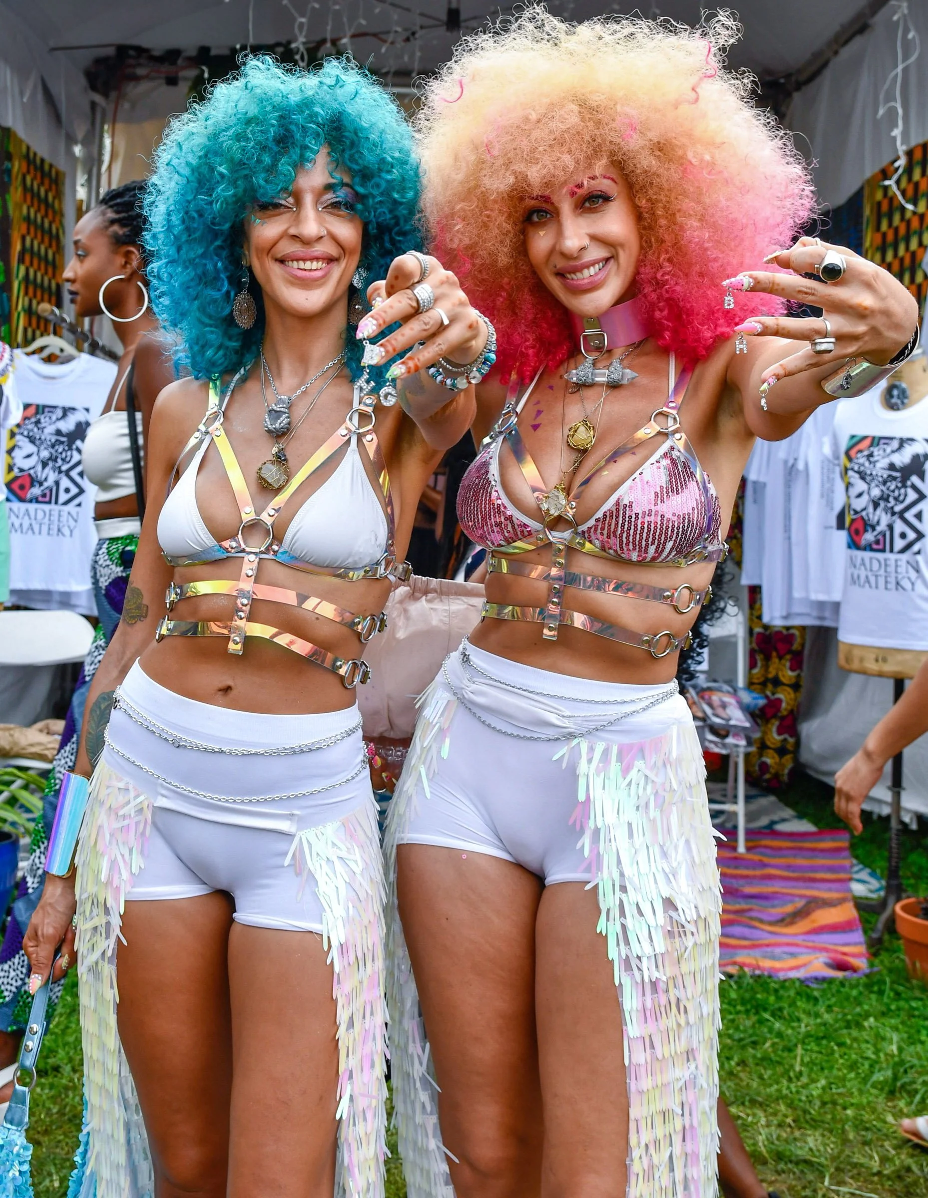Two women with colorful curly hair, wearing shiny harnesses and white shorts, are smiling and posing at a lively outdoor event with other people and booths in the background.