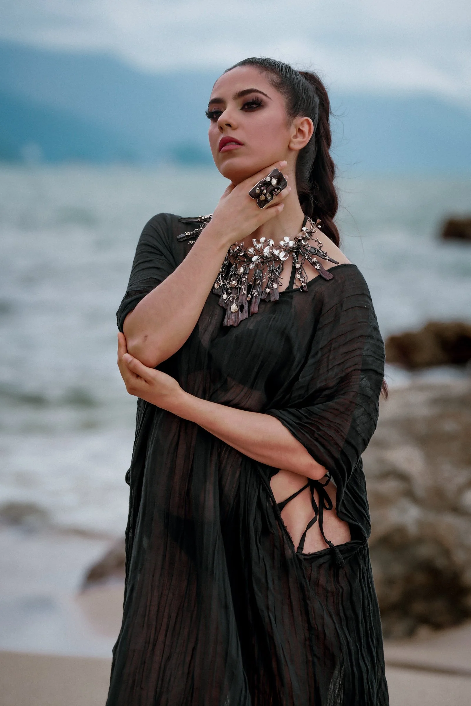 A woman with dark hair styled in a high ponytail, wearing bold makeup, a statement necklace, and a black dress with revealing side details, stands on a beach with the ocean and cloudy sky in the background.