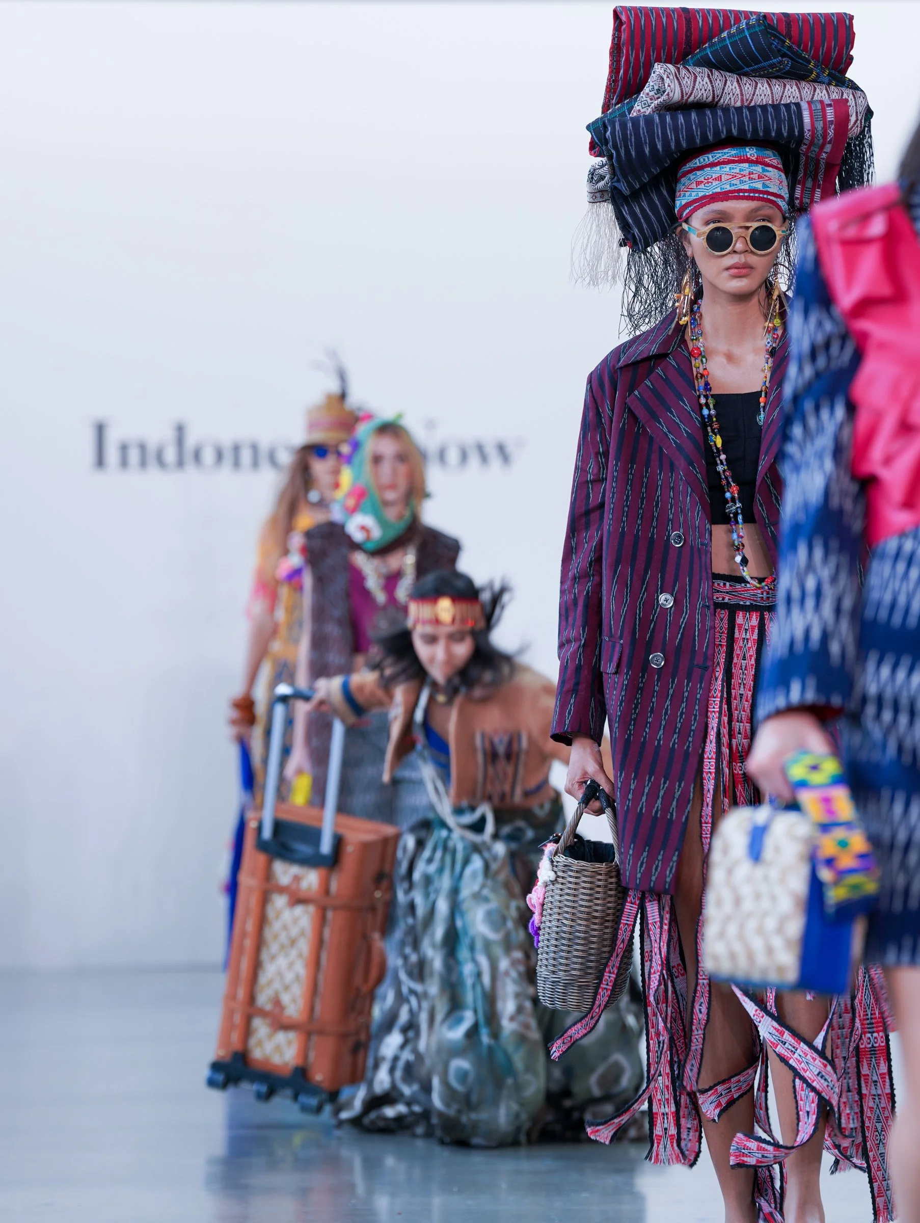 Fashion show with models dressed in colorful, traditional woven fabrics from Indonesia. The models are accessorized with sunglasses, jewelry, and carry bags, walking on a runway with a white background and the word "Indonesia" visible.