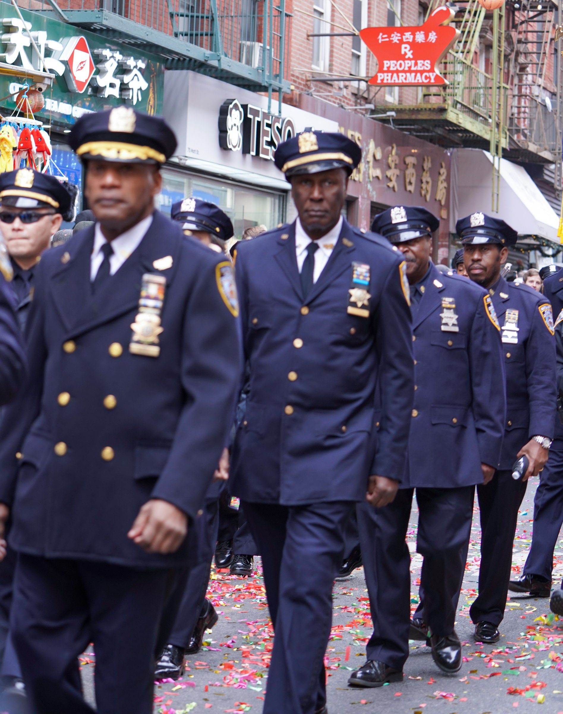 A group of police officers in uniform walking in a parade on a city street decorated with colorful confetti. There are storefronts and signs in the background.