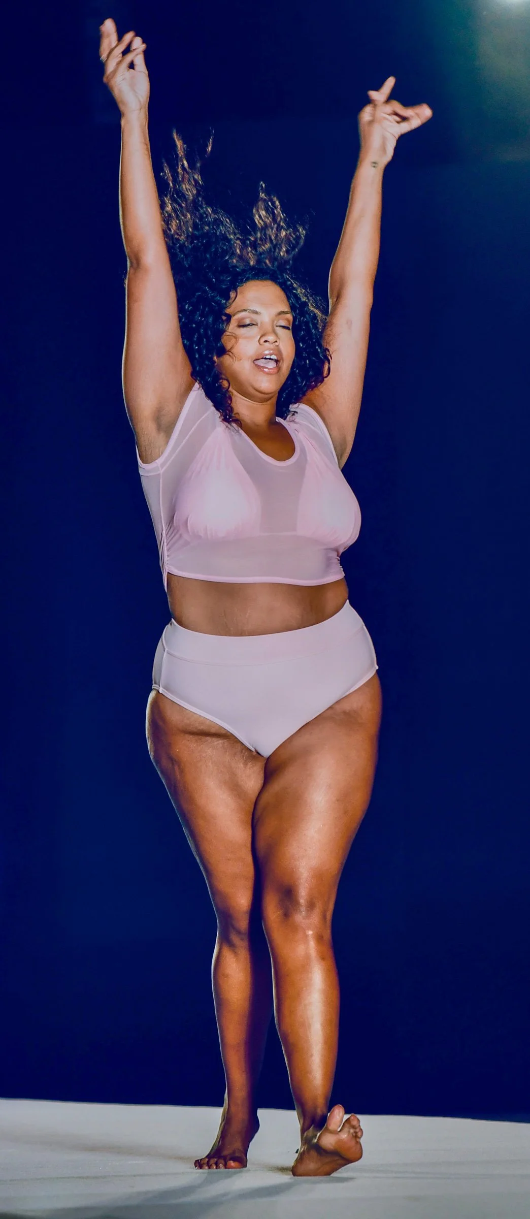 A woman with curly hair, wearing a pink transparent top and white high-waisted underwear, raising her arms with closed eyes against a dark blue background. Sports Illustrated Swimsuit Show at W Hotel South Beach.