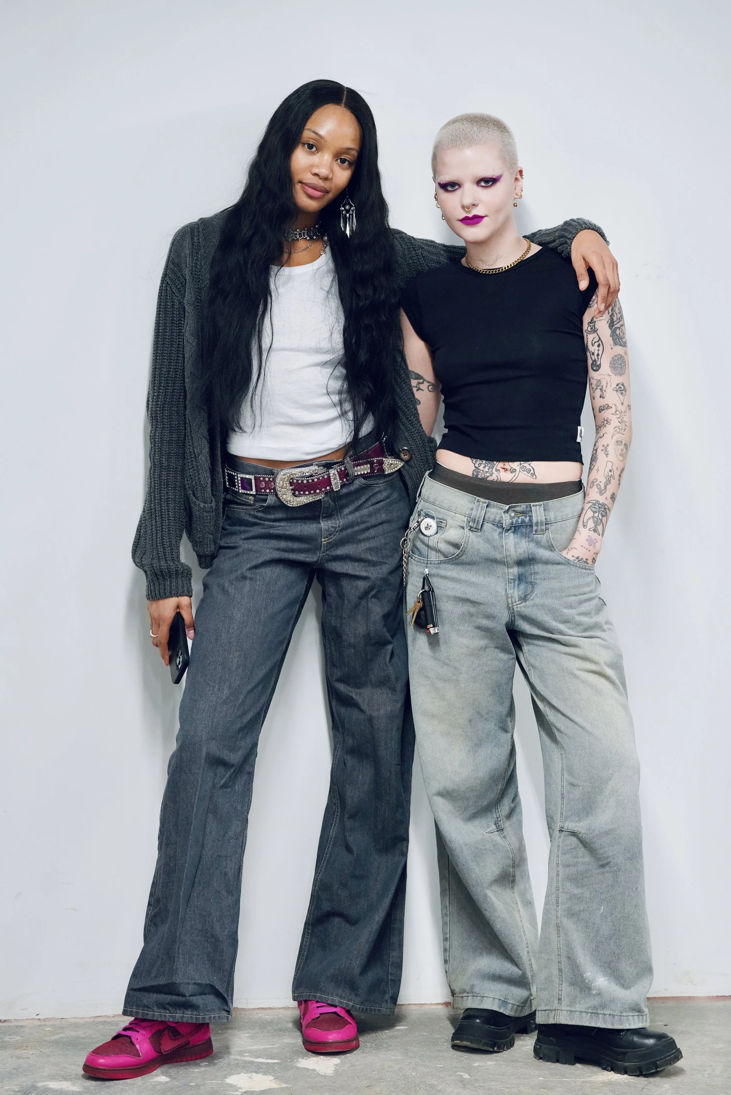 Two women standing close together against a plain background, one with long black hair wearing a gray cardigan and dark jeans, the other with short blonde hair, tattoos, and dark makeup, wearing a black sleeveless top and baggy jeans.