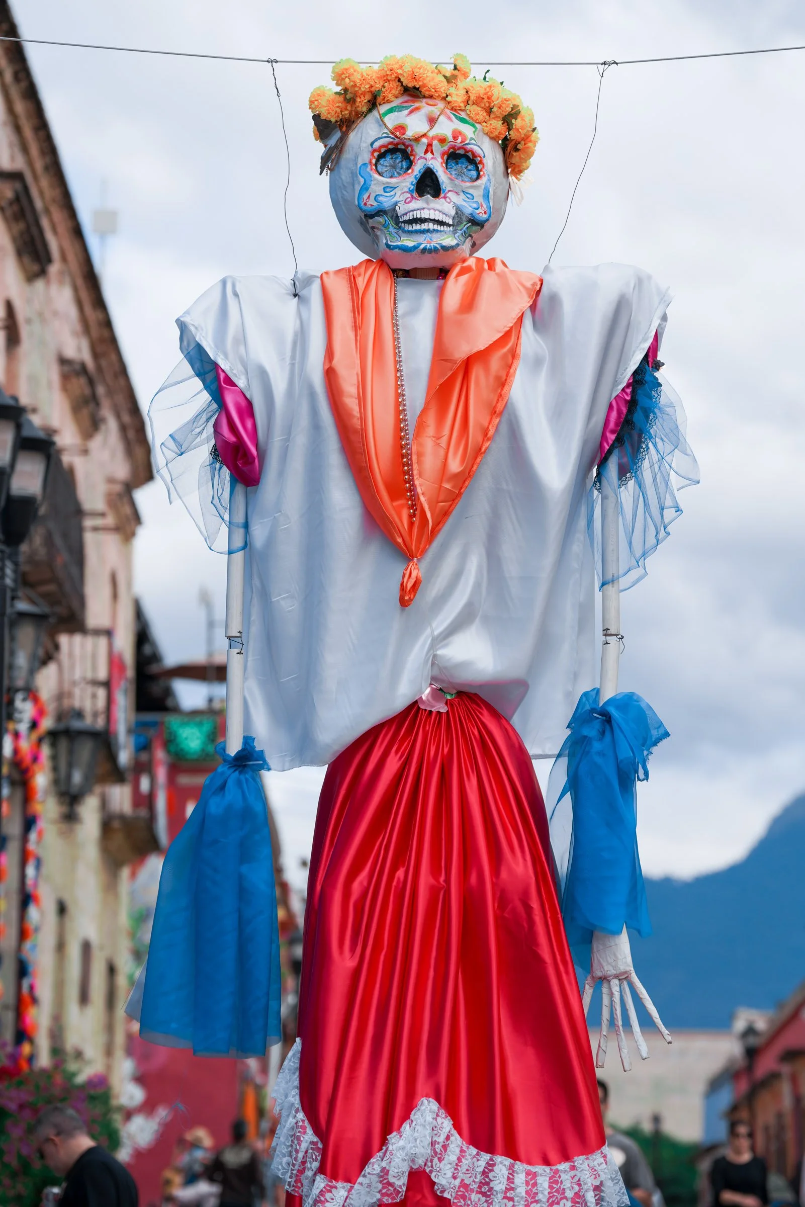 A large marionette with a skull face, decorated with colorful patterns, wearing a floral crown and a dress with bright colors, is being operated in a street festival with buildings and people in the background.