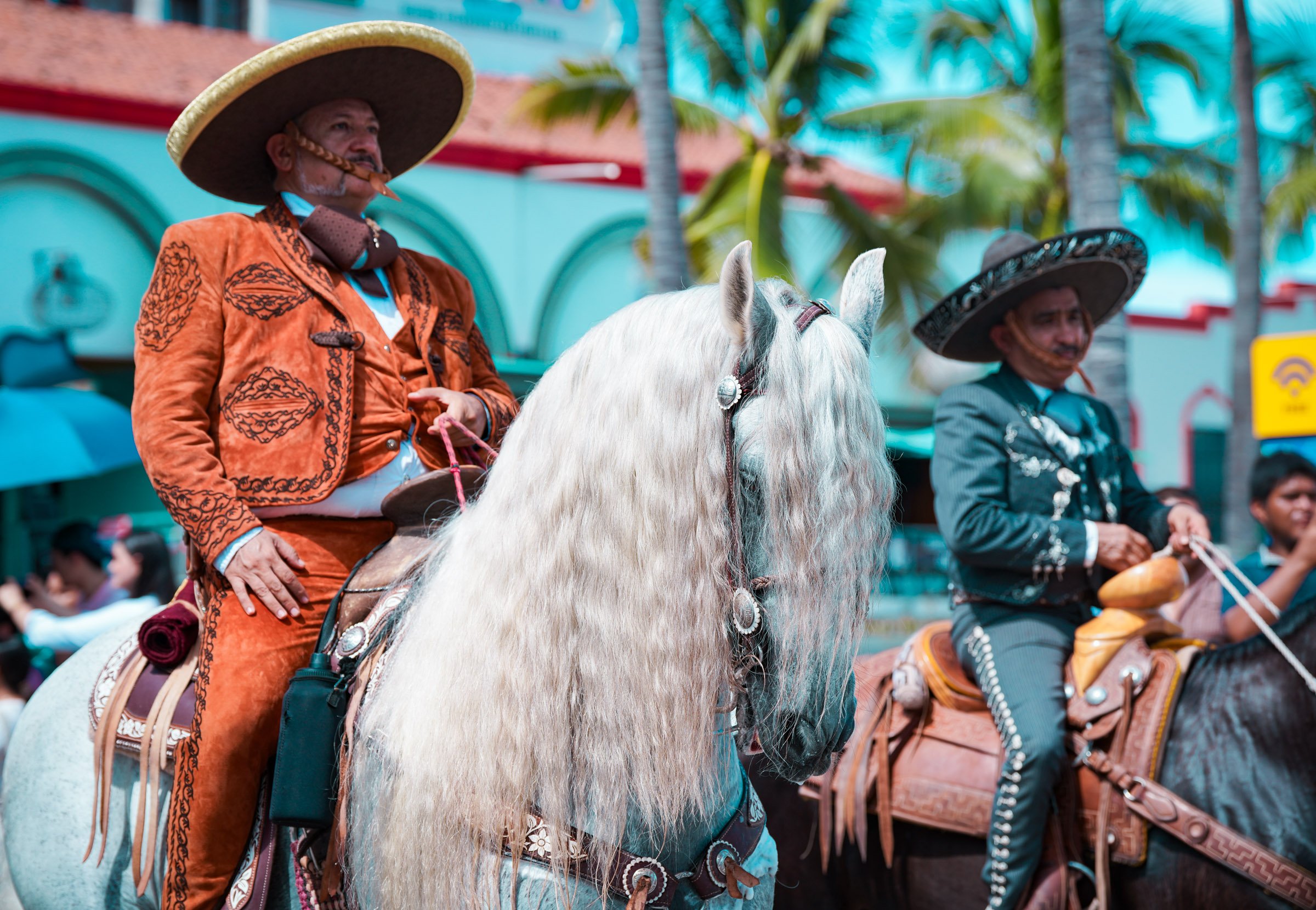 Two men dressed in traditional Mexican attire riding decorated horses during a lively parade or celebration. The scene is bright and colorful, with palm trees in the background.
