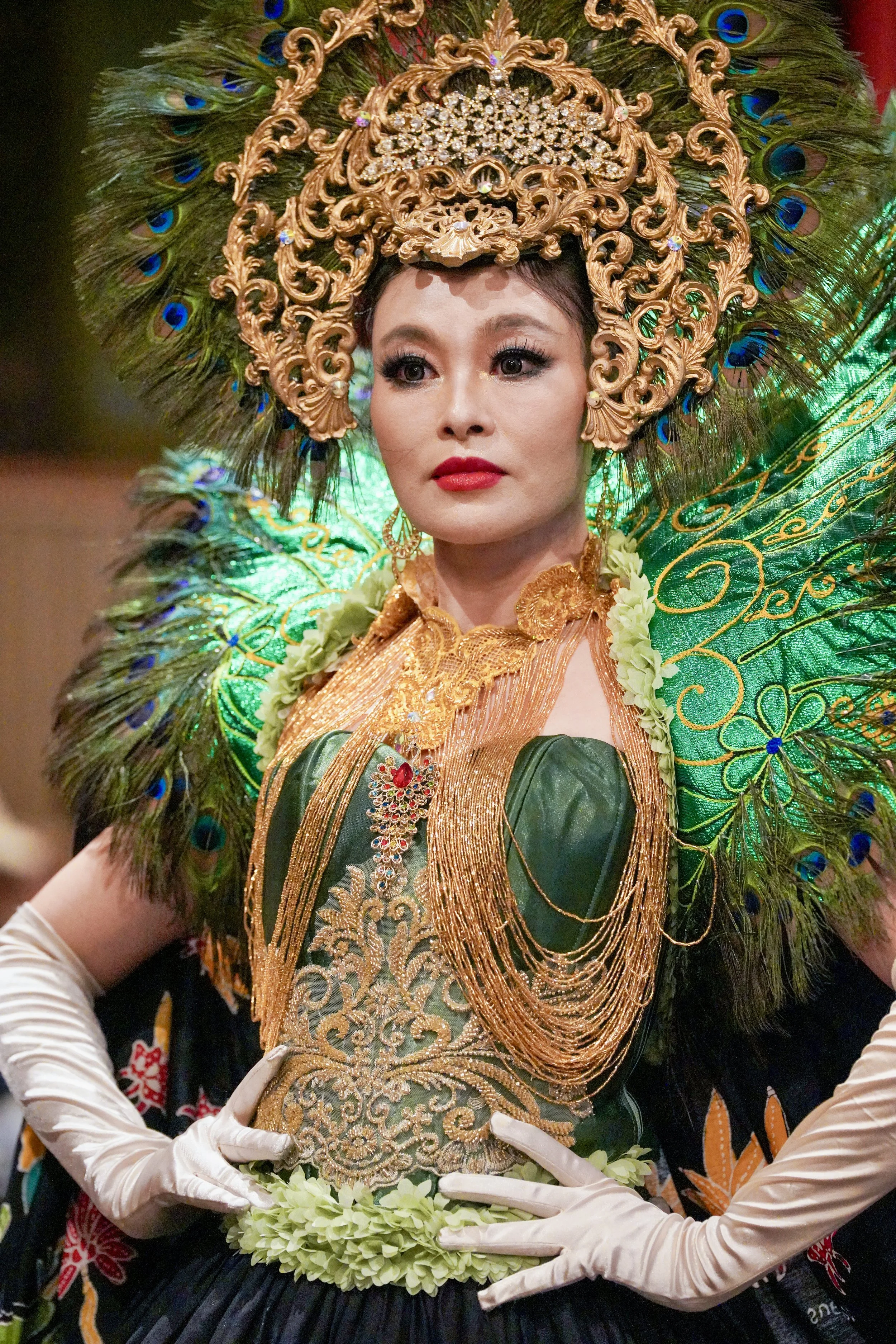 Woman wearing ornate traditional costume with large peacock feather headdress and gold embroidery, holding a green floral necklace. Asian NY Fashion Week during NYFW.