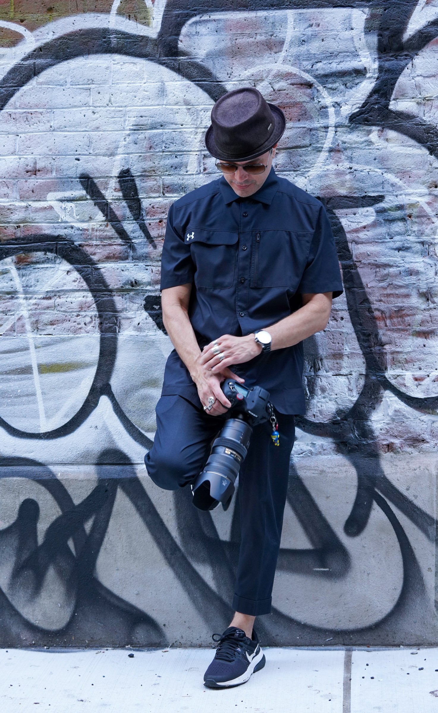 Person in black clothing, wearing a hat and sunglasses, leaning against a graffiti-covered brick wall, holding a professional camera with a long telephoto lens.