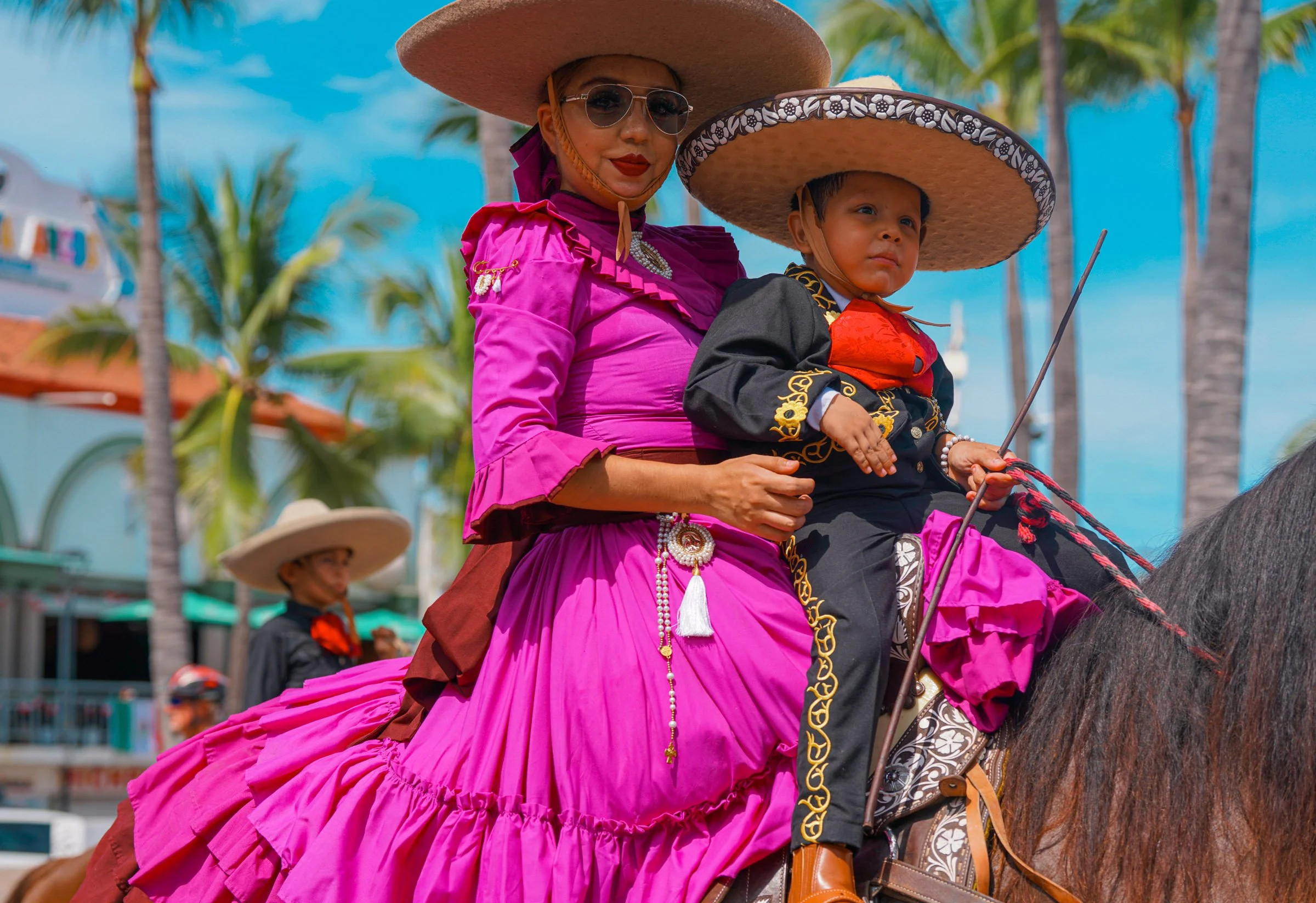 A woman dressed in a vibrant pink traditional outfit and wearing a large straw hat, sunglasses, and red lipstick, sitting on a horse. A young boy in a black and red outfit is seated in front of her, also wearing a large straw hat, with a background o