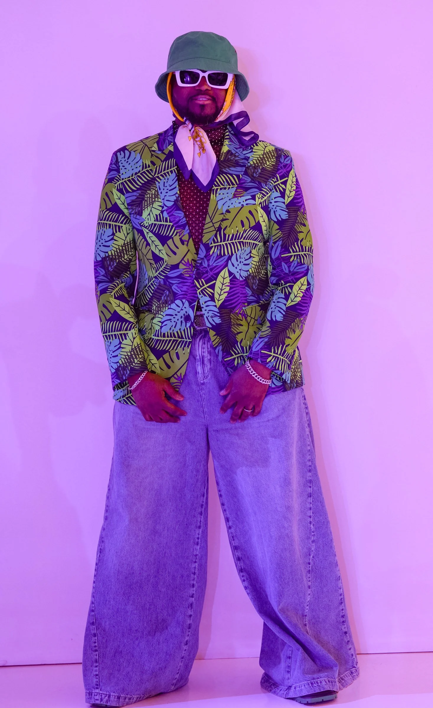 Person wearing a green bucket hat, white sunglasses, a patterned blazer with leaves, a white and purple neck scarf, loose jeans, and jewelry, standing against a purple wall.