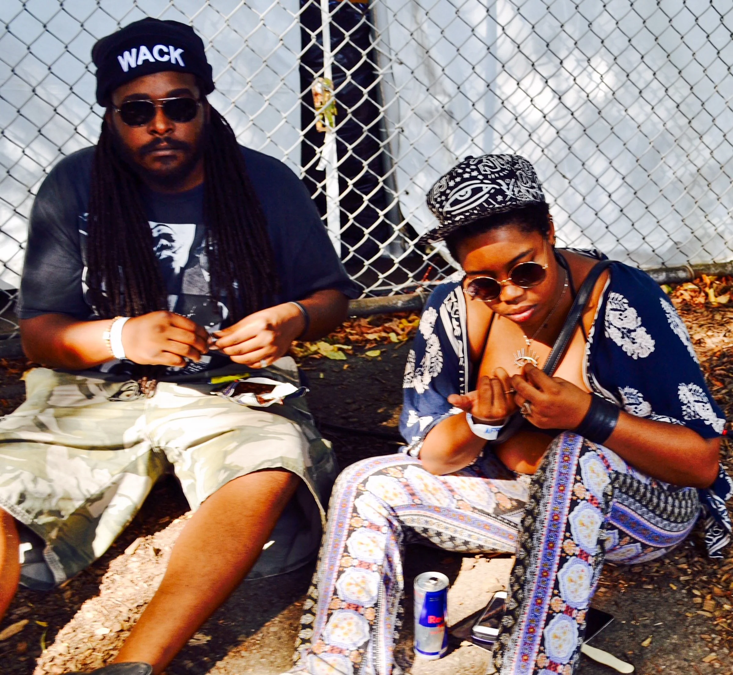 Two individuals sitting outdoors near a chain-link fence. The man on the left has long dreadlocks, sunglasses, a black beanie with the word "WACK" on it, and wears dark clothing. The woman on the right has short hair, round sunglasses, and is dressed
