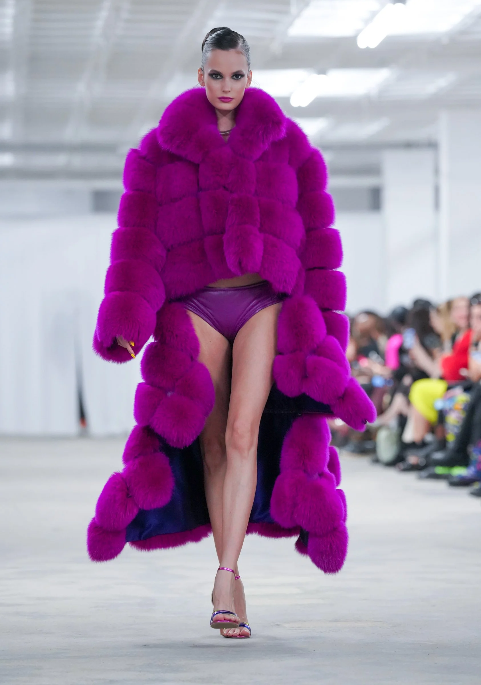 Model walking on runway wearing a vibrant purple fur coat with matching purple underwear, high heels, and slicked-back hair in an indoor fashion show.