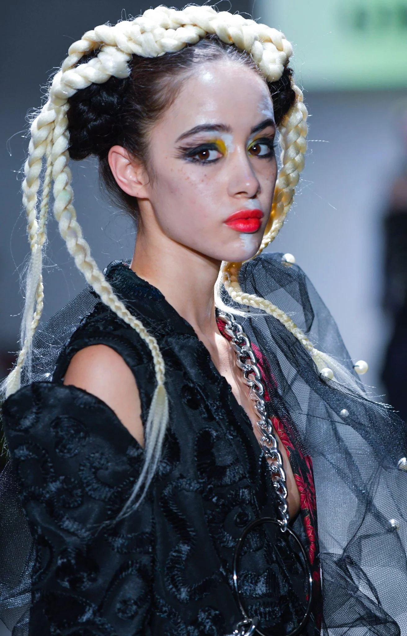 A woman with dramatic makeup and platinum blonde braids wearing a black velvet jacket with mesh and pearl accents, and accessorized with a large chain necklace and a black circular pendant.