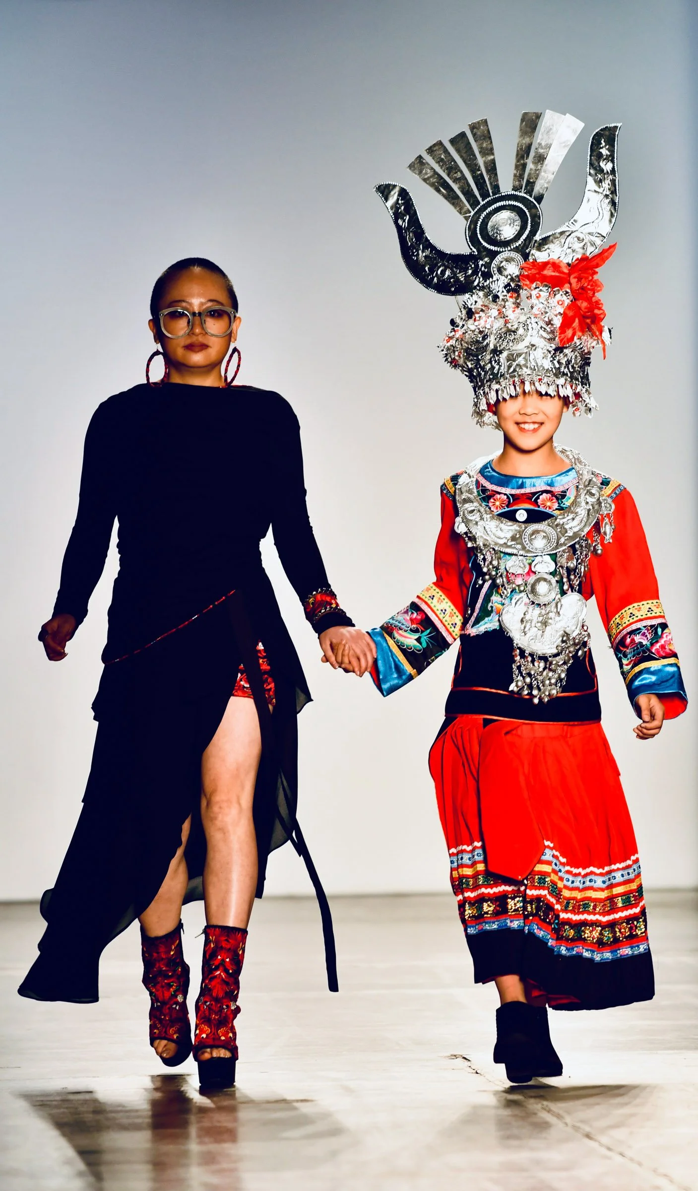 Two women walking hand in hand on a runway, wearing traditional and modern fashion. The woman on the left is dressed in a black outfit with a high slit, red floral-patterned boots, and sunglasses. The woman on the right is wearing a vibrant, ornate t