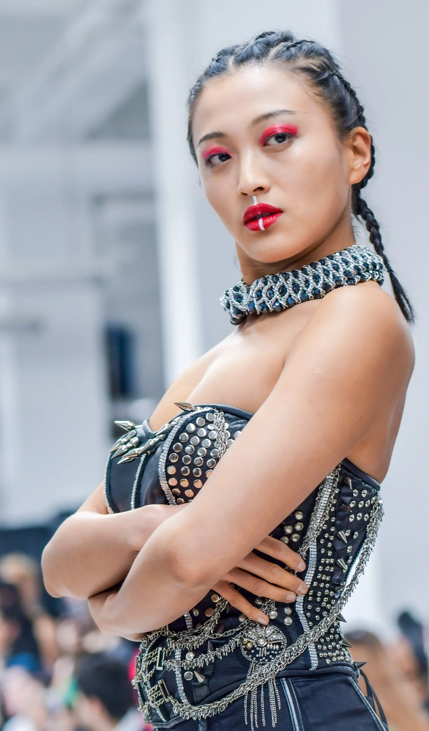 A young woman with braided hair and red makeup, wearing a studded leather outfit with chains, crosses her arms and looks confidently at the camera.