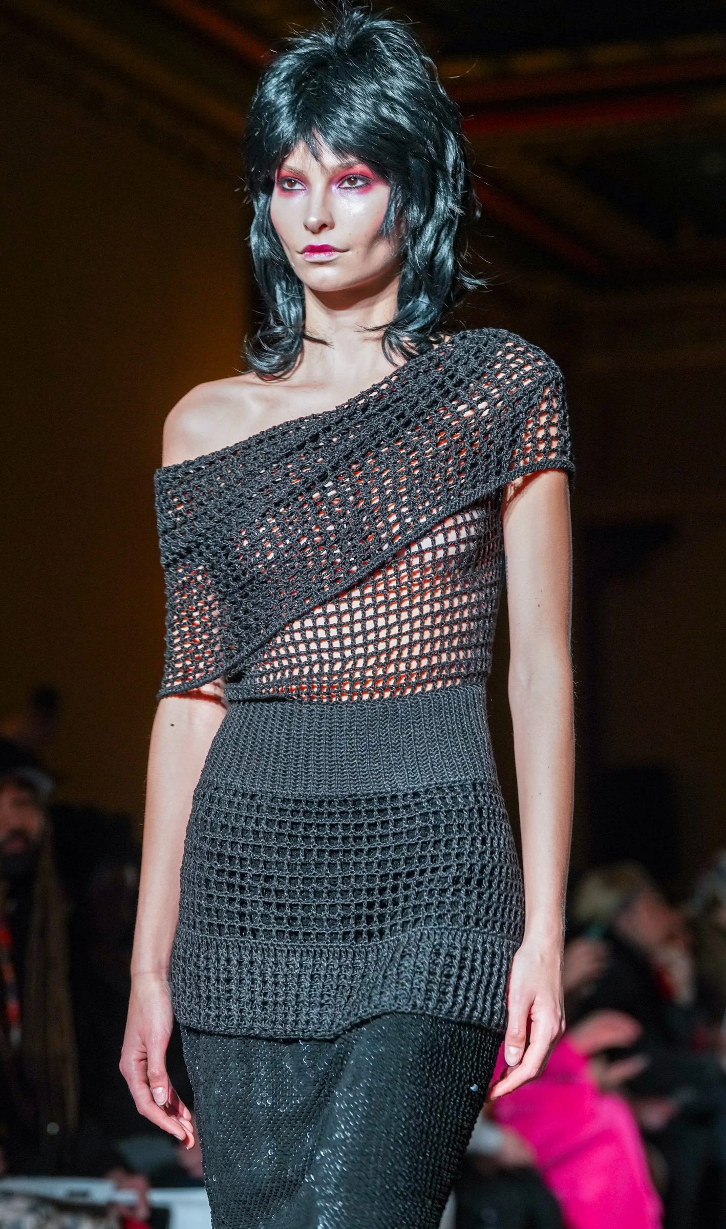 Model walking on runway wearing a black crochet off-shoulder top with red eyeshadow and black hair.