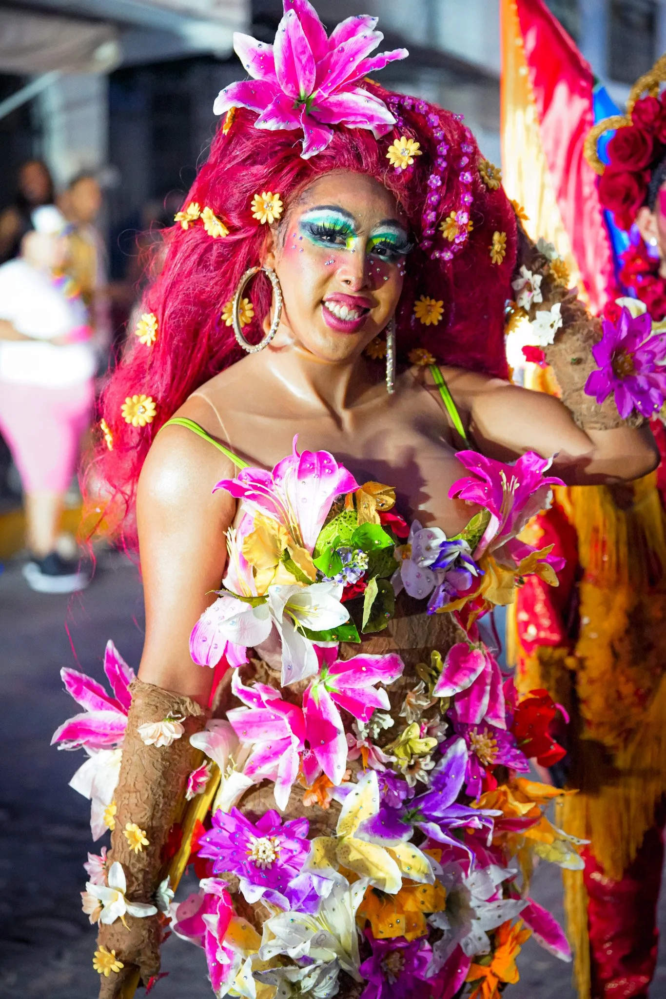A person dressed in colorful floral costume with pink hair, large hoop earrings, vibrant makeup, and numerous flowers adorning their hair and body, participating in a parade or festival.