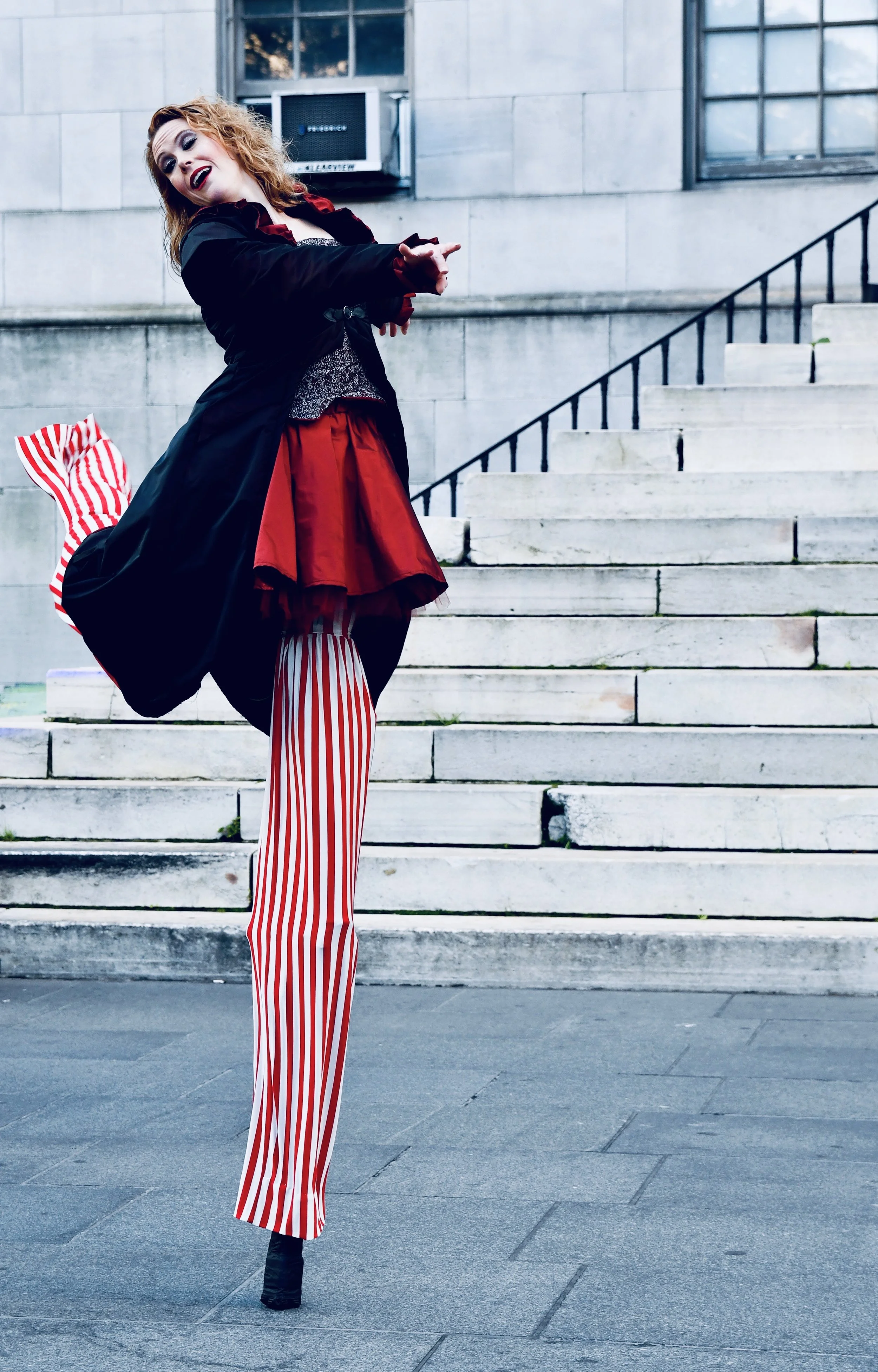 A person with curly hair dressed in a striped red and white pant, a black jacket, and a red skirt, standing outdoors on concrete steps, pointing and singing with a lively expression.